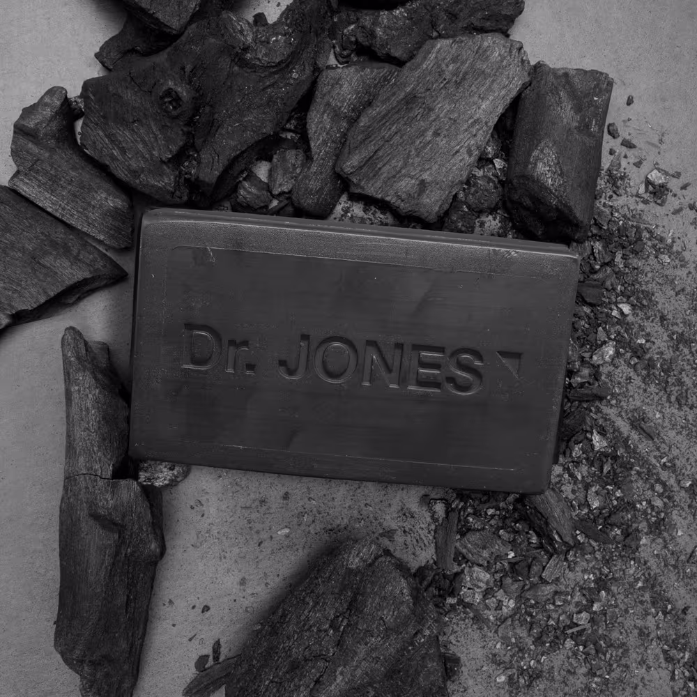 A imagem mostra pedaços de carvão e no meio um sabonete retangular com o nome "Dr. Jones" gravado. A imagem remete a um ambiente industrial.