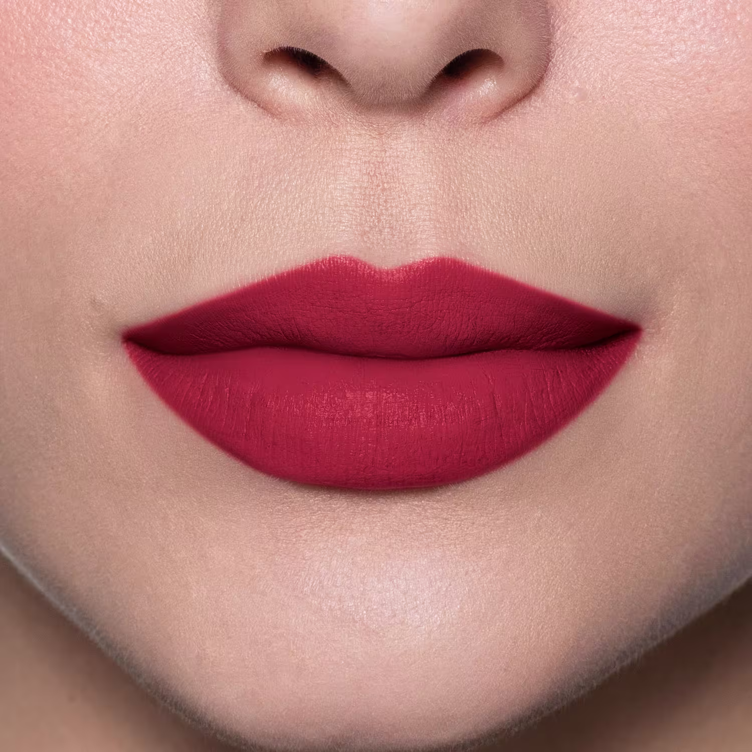 A imagem mostra um close do batom aplicado nos lábios de uma pessoa branca. Os lábios parecem macios e bem delineados, com um acabamento discretamente brilhante. O tom vermelho transmite uma aparência sofisticada e elegante.