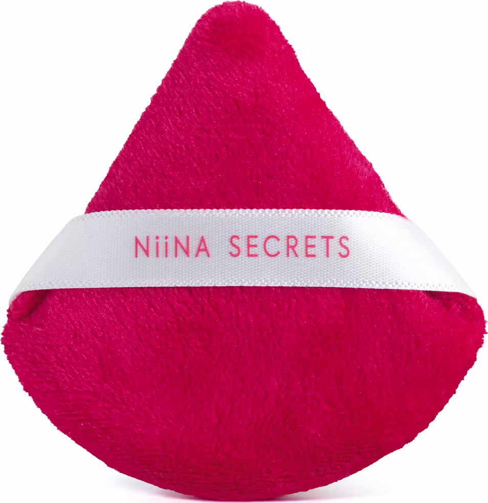 A imagem mostra uma esponja de maquiagem triangular de cor vermelha com uma fita branca na parte central, com  o nome da linha, Niina Secrets.