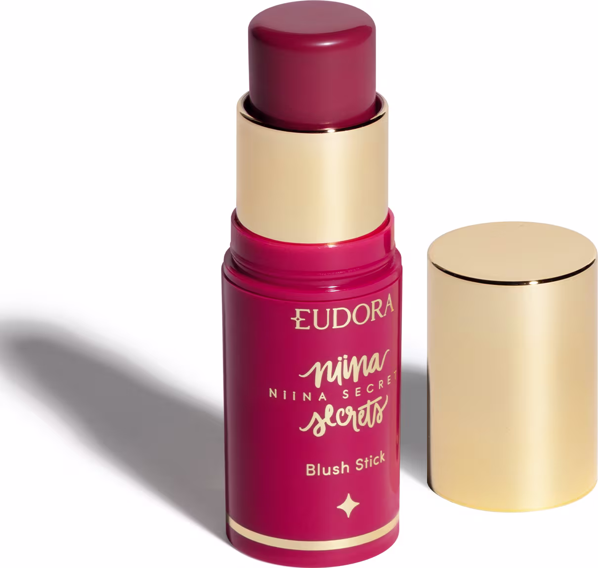 O produto apresentado é um blush stick da linha Niina Secrets da marca Eudora. Possui uma embalagem rosa com detalhes dourados, incluindo o nome da marca e da linha. O produto tem formato cilíndrico e tampa dourada na parte superior.