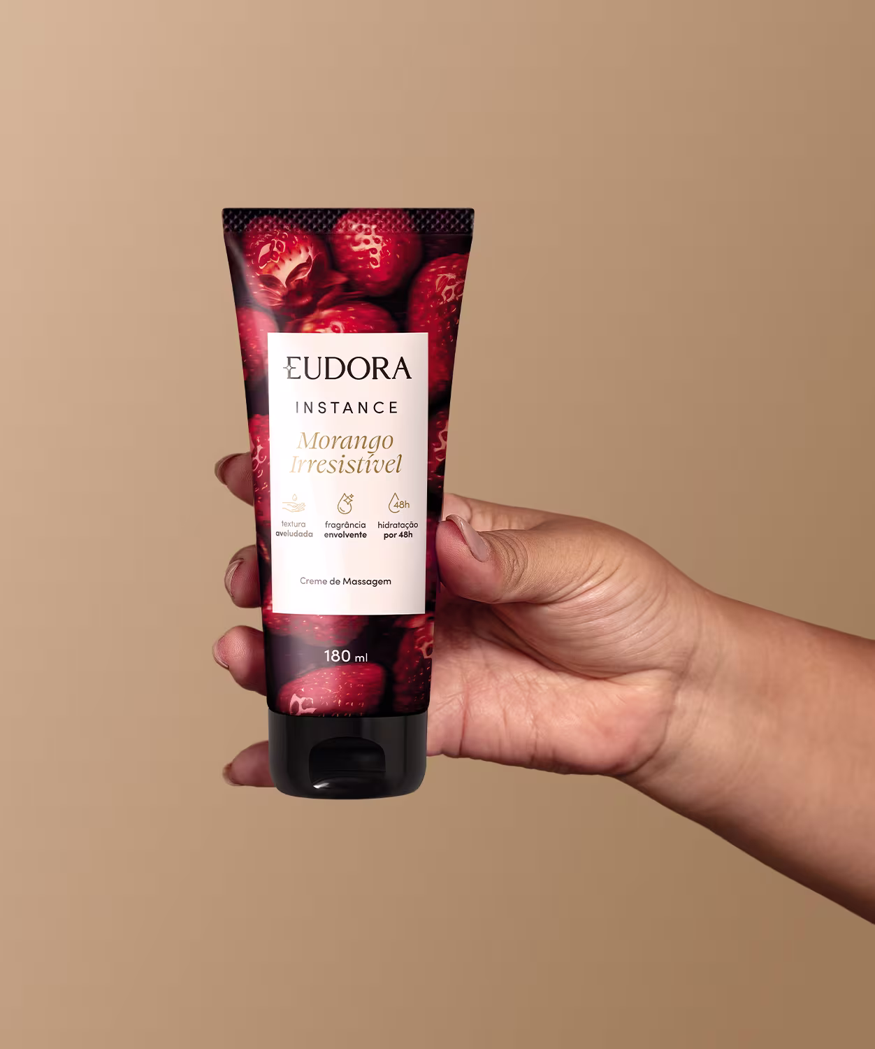 O produto mostrado na imagem é um creme de massagem da linha Instance Morango Irresistível. A embalagem é um tubo plástico preto com uma imagem de morangos vermelhos brilhantes na parte frontal, que é segurado por uma mão feminina.