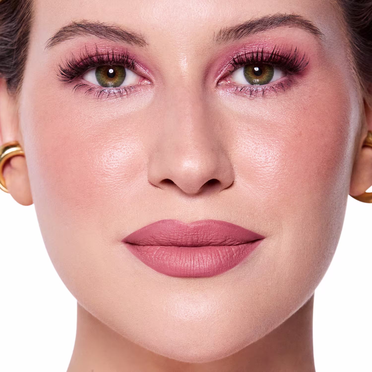 Rosto da Niina em foco, com maquiagem rosada e batom rosa claro semi-matte. Olhos verdes destacados por cílios longos e sobrancelhas bem definidas. Fundo claro e iluminado, sugerindo um ambiente de beleza ou cosmética.
