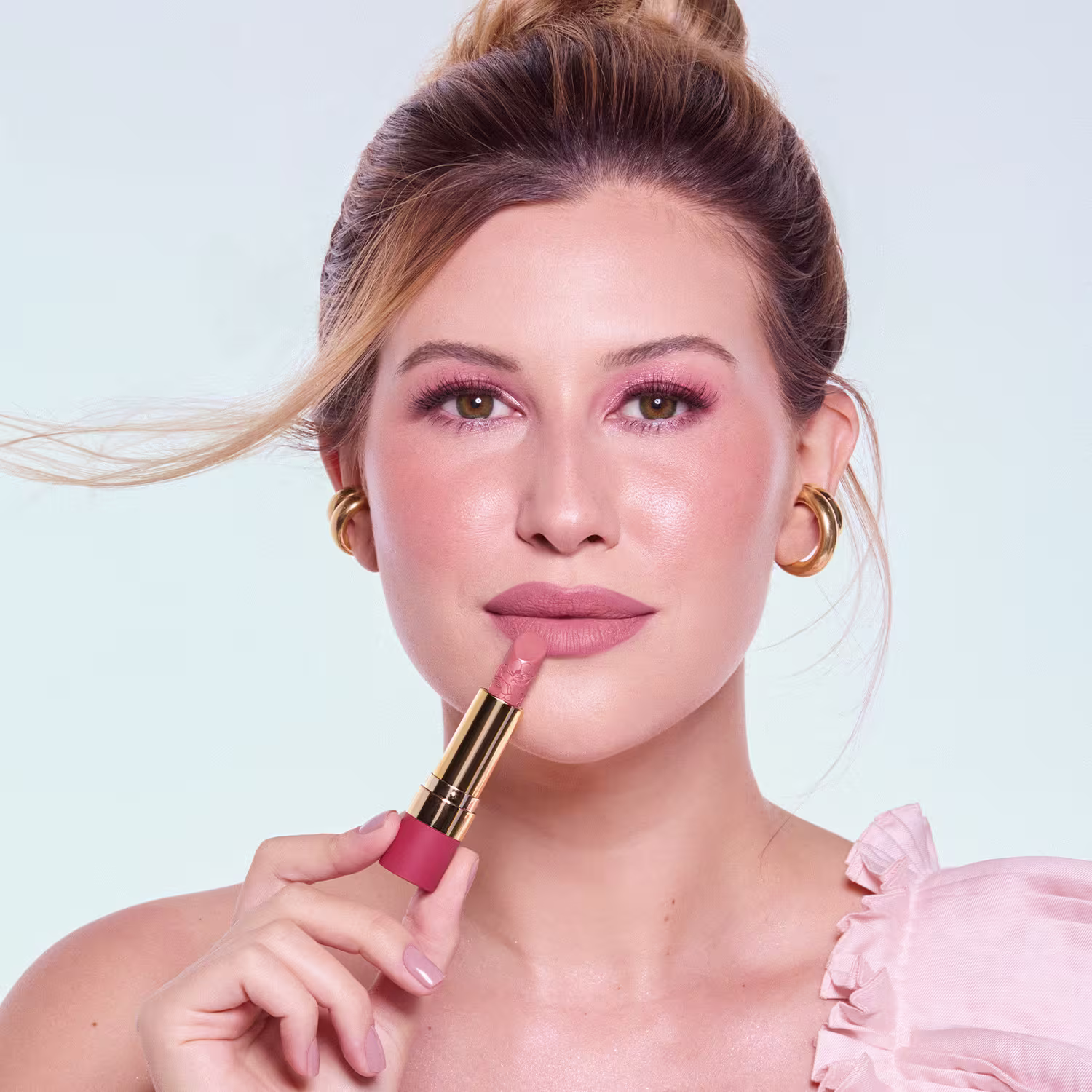 Imagem da Niina segurando o batom Rosé Pétala aberto, próximo a boca. Seu rosto está em primeiro plano, com maquiagem suave e cabelo preso. O ambiente sugere um contexto de beleza e cuidados pessoais.