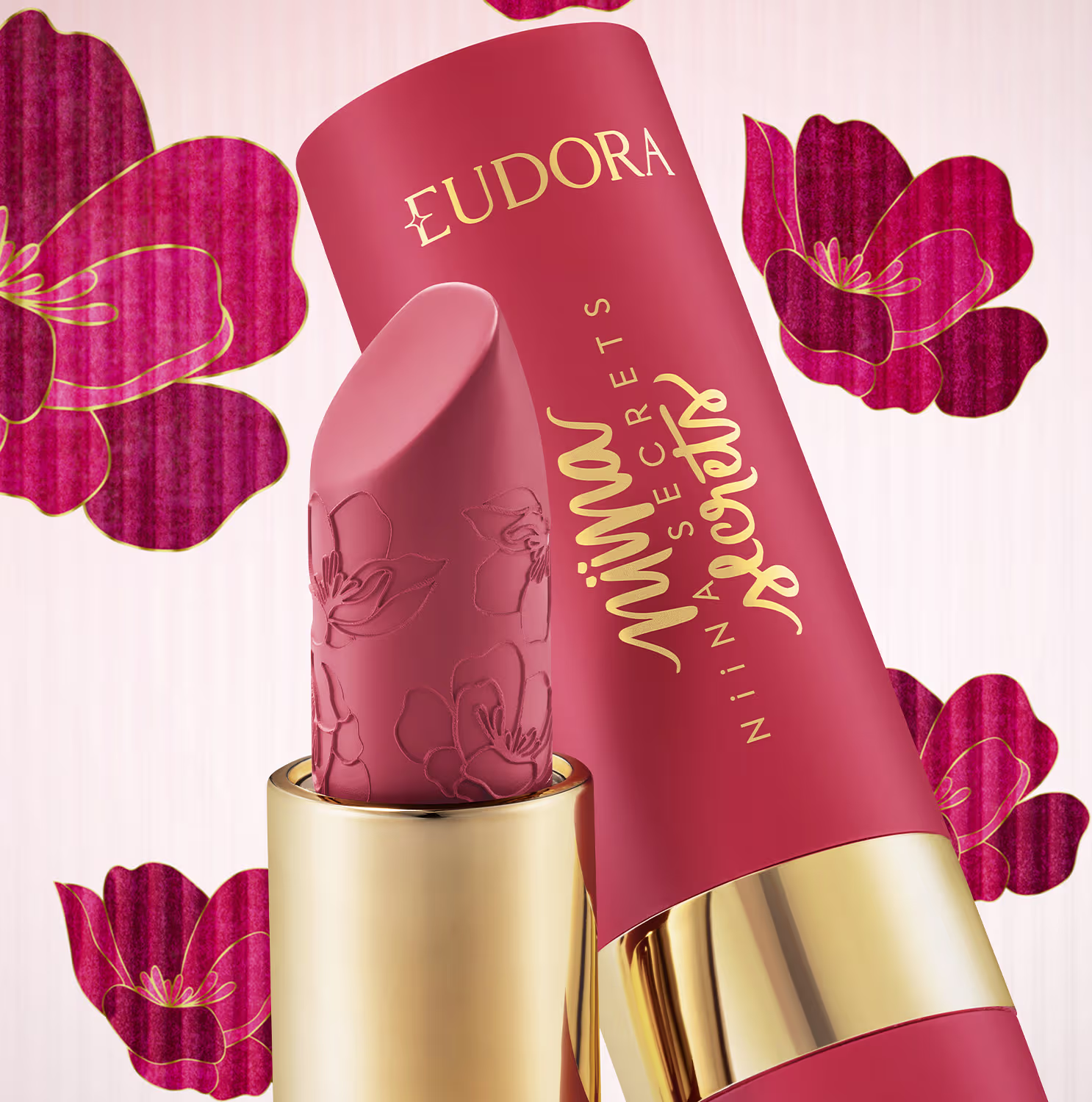 Recipiente de batom da marca Eudora em formato cilíndrico com decoração floral em relevo. Embalagem rosada com detalhes dourados. Fundo rosa claro com flores vermelhas grandes em estilo artístico.