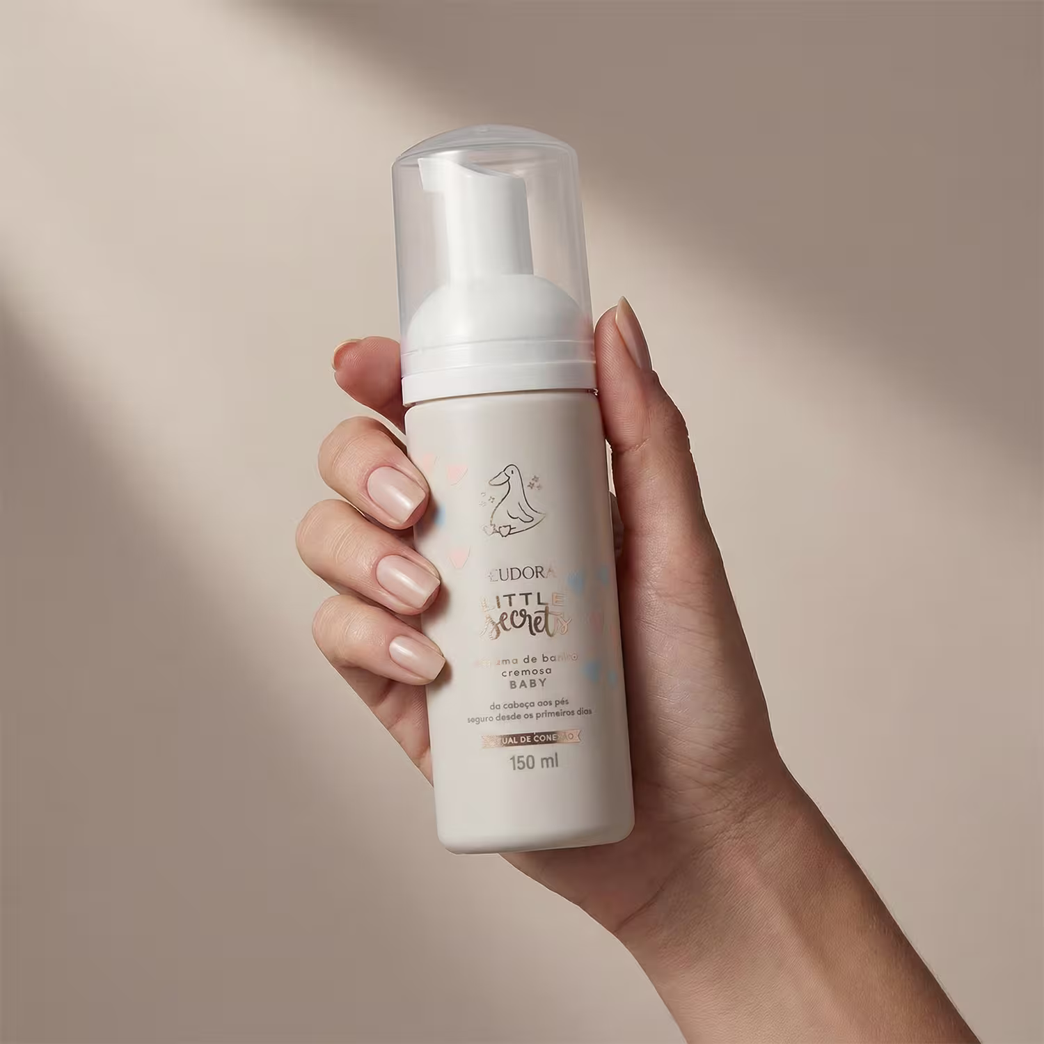 Produto de cuidados para bebê da marca Little Secrets, em embalagem branca com detalhes em azul claro, sendo segurado por mãos femininas com unhas curtas e esmaltadas em tons claros, em um ambiente com iluminação suave e tons neutros.