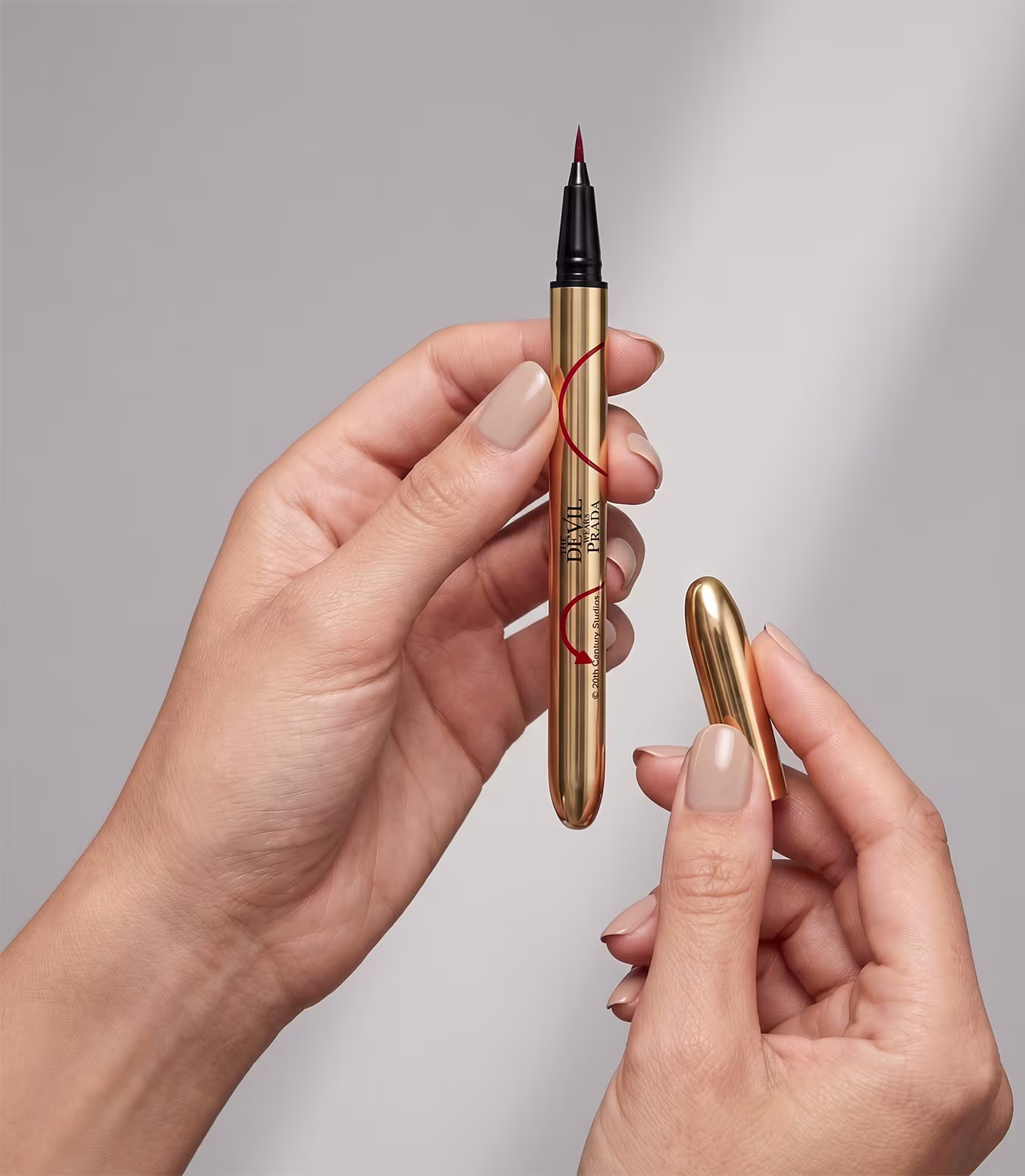 Caneta delineadora dourada com ponta fina, com rótulo "The Devil Wears Prada" em letras pretas, sendo segurada por mãos femininas com unhas curtas e pintadas.
