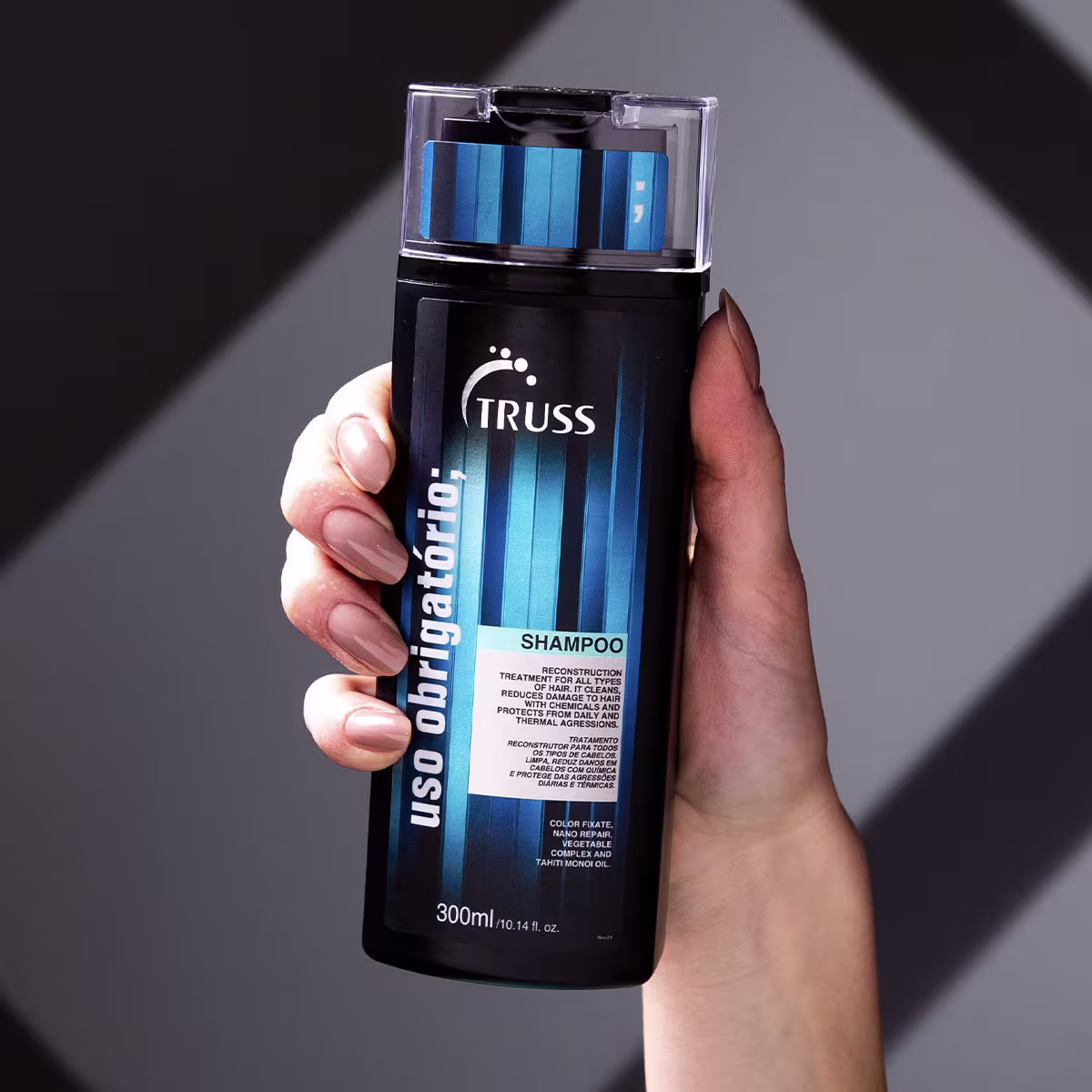 A imagem mostra uma mão segurando um frasco de shampoo da marca Truss. O frasco é preto com detalhes em azul e tem uma tampa transparente. Além disso, a embalagem mostra benefícios como redução de danos e proteção.