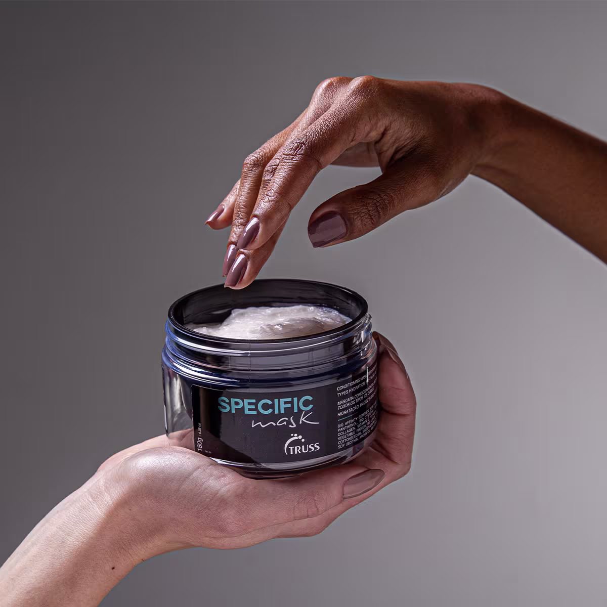 A imagem mostra uma embalagem de plástico contendo uma máscara capilar da marca Truss: Specific Mask. Uma mão feminina segura o frasco e está com o dedo indicador dentro dele, provavelmente aplicando ou retirando um pouco do produto cremoso branco.
