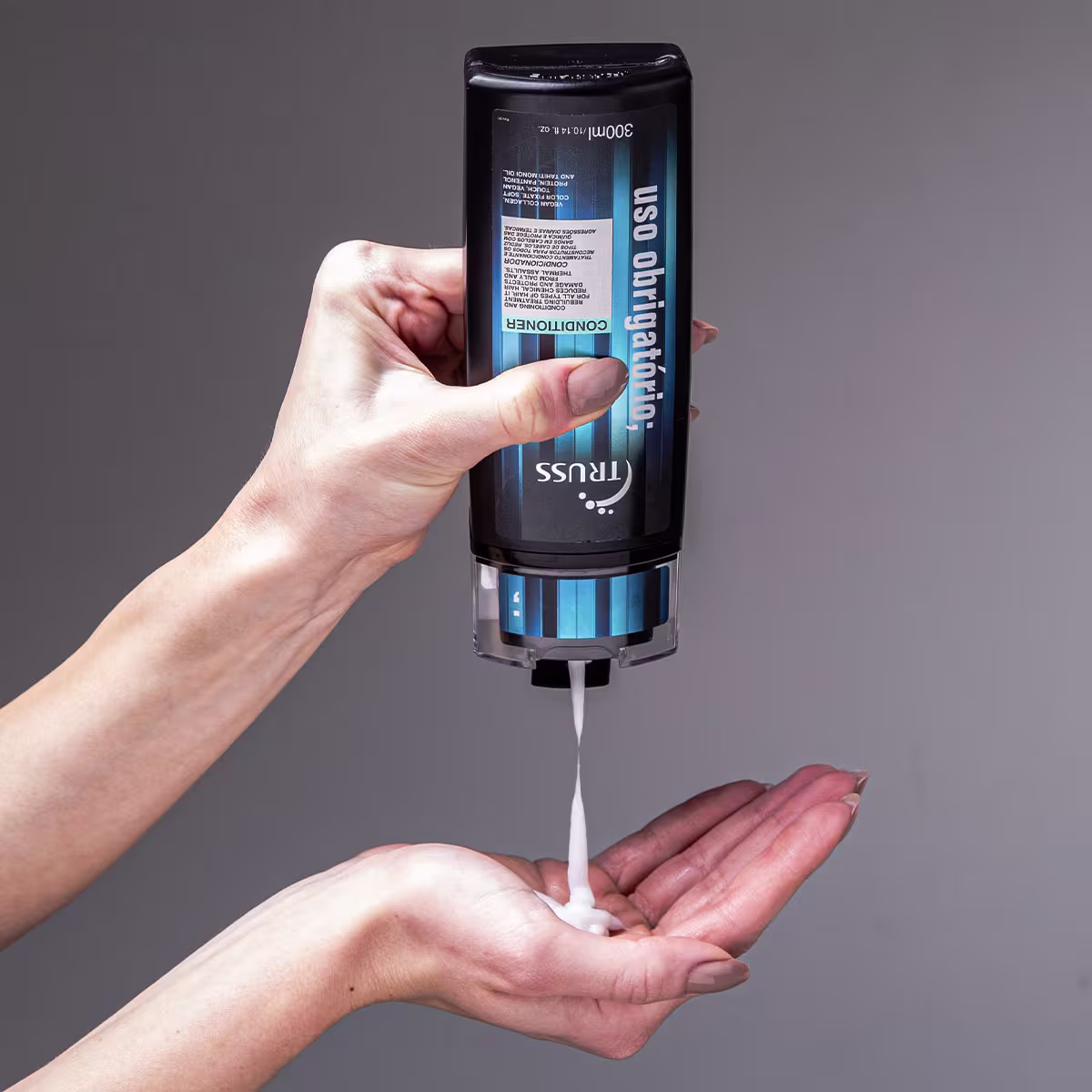 A imagem mostra uma mão segurando um frasco grande de condicionador de cabelos da marca "Truss", da linha "Uso Obrigatório. O frasco é preto com detalhes em azul. Um líquido branco está sendo dispensado para a palma da outra mão, aparentando ser cremoso.