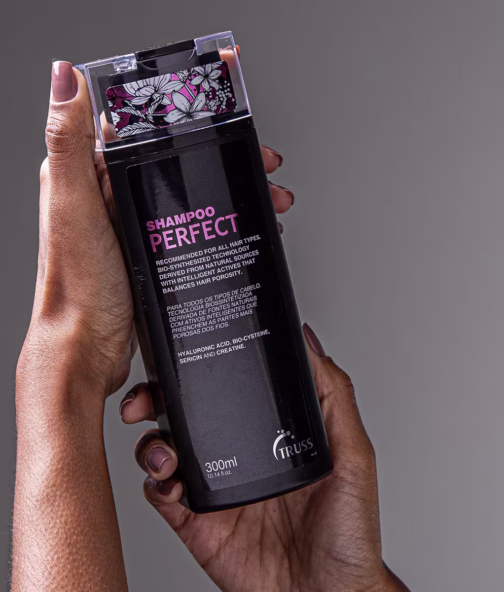 A imagem mostra uma mão segurando um frasco grande de shampoo da marca Truss. O shampoo é chamado "Perfect" e, de acordo com a embalagem em português, é recomendado para todos os tipos de cabelo. O frasco é preto com detalhes florais na parte superior.