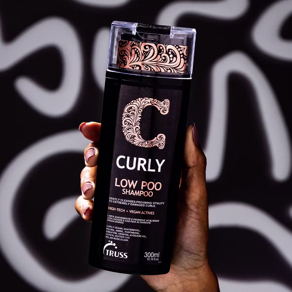 A imagem mostra um frasco de 300ml de shampoo "Curly" da Truss, para cabelos cacheados e ondulados. O frasco escuro é segurado por uma mão, destacando seu design floral elegante e a descrição "Deep Treatment for Curly and Wavy Hair".