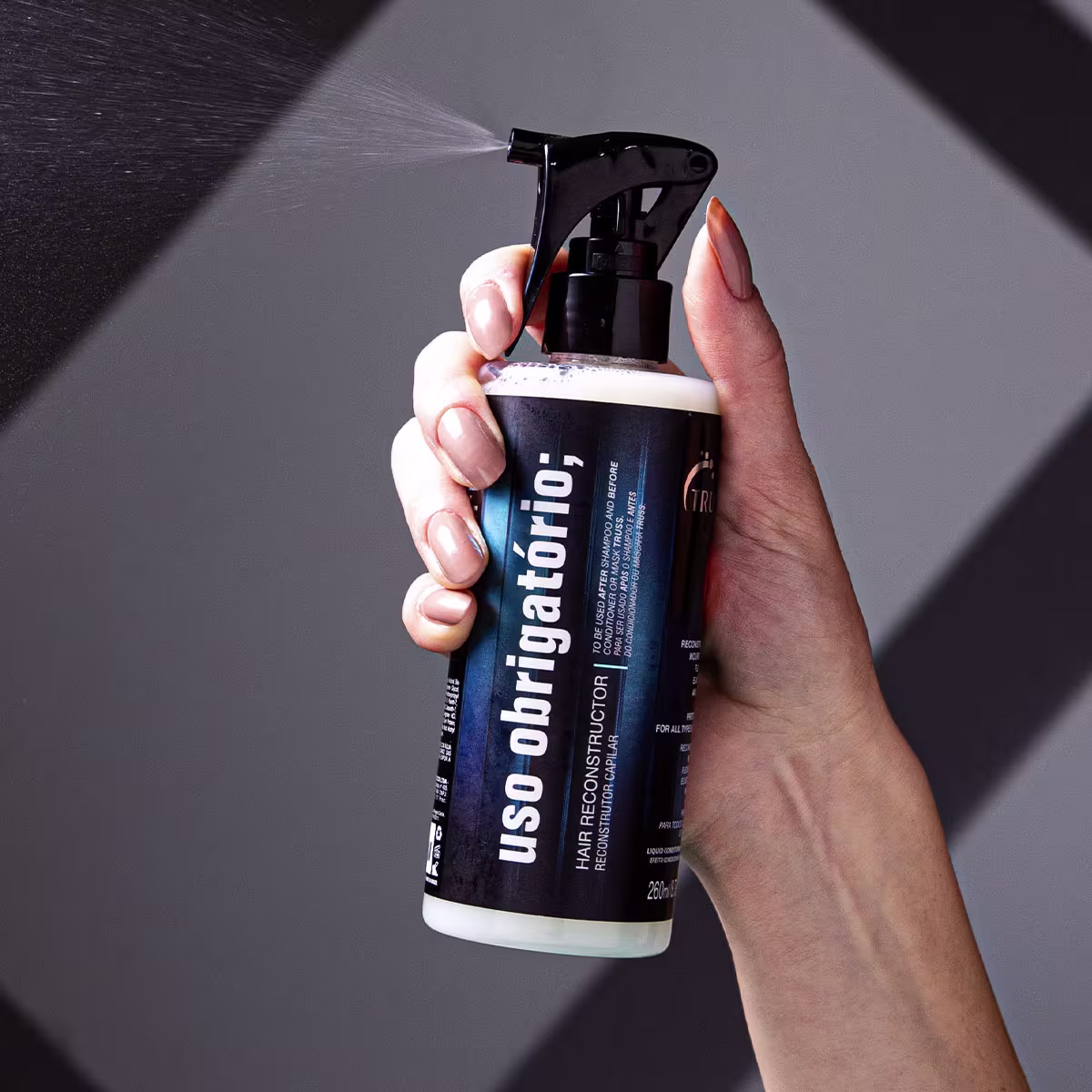 A imagem mostra uma mão segurando um produto da marca Truss. O frasco é azul e preto, e está escrito "Uso Obrigatório" na embalagem, indicando que se trata de um reconstrutor capilar.