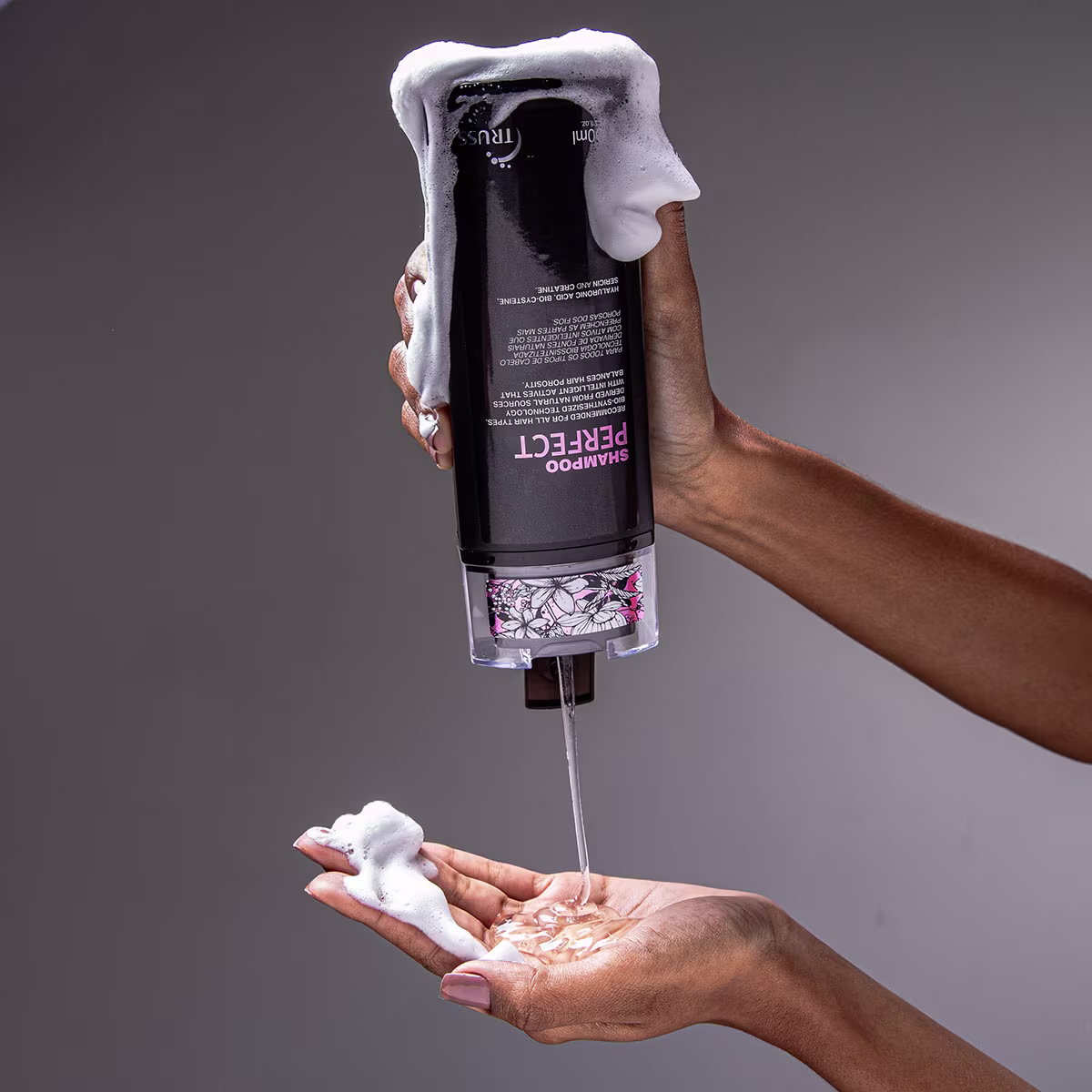 Na imagem um par de mãos segura uma garrafa preta, de shampoo, da qual sai um líquido formando uma espuma branca na outra mão.