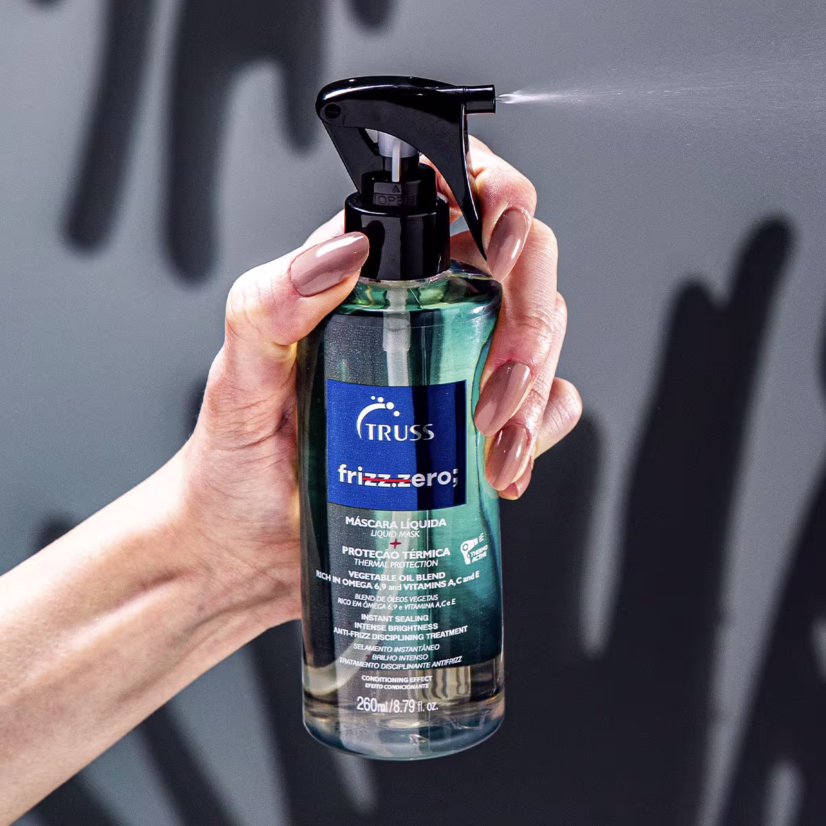 A imagem mostra uma mão feminina segurando a máscara líquida Frizz Zero da marca TRUSS: embalagem plástica transparente de 260ml com rótulo azul e borrifador, além das descrições do produto em português e inglês.