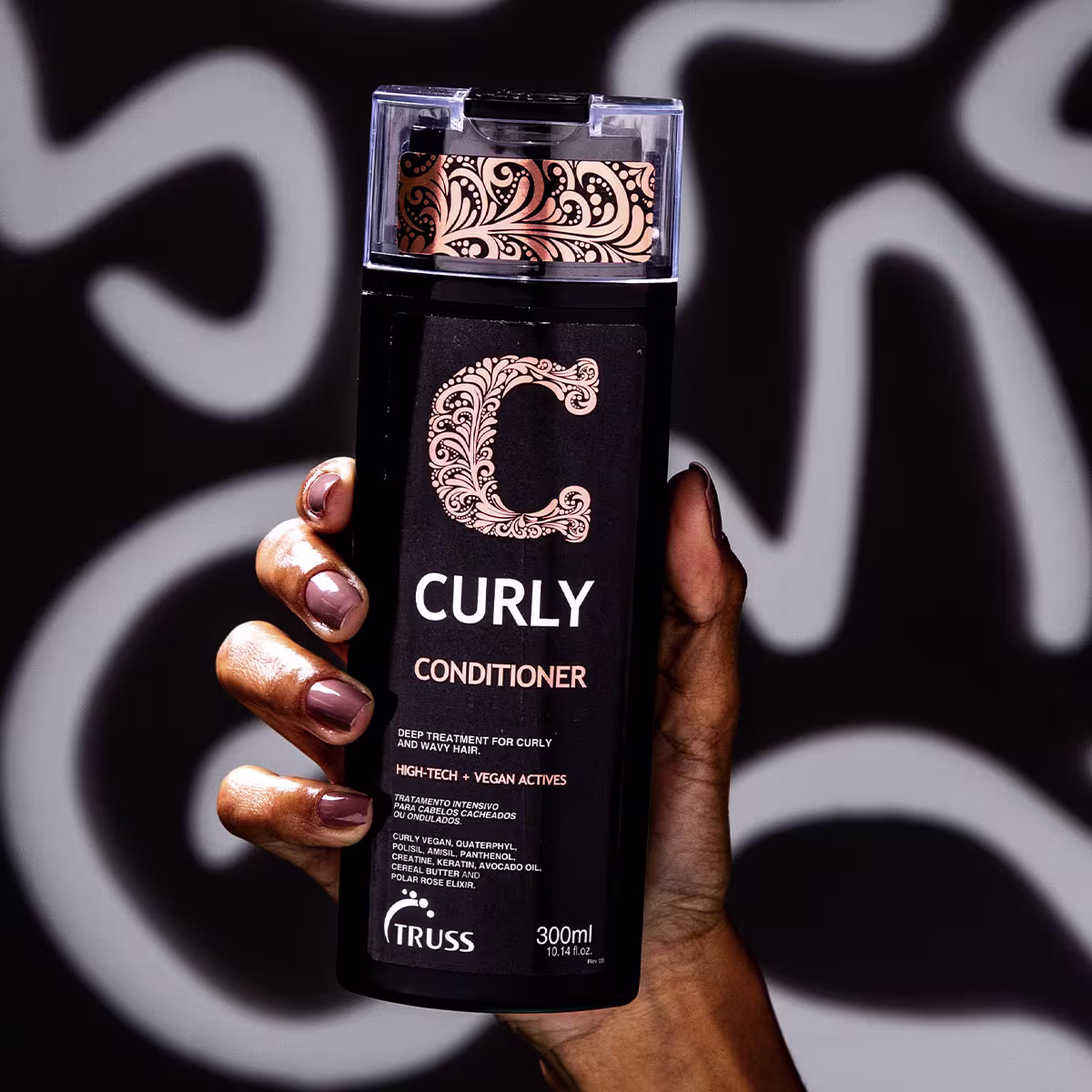A imagem mostra uma mão feminina segurando a embalagem do Condicionador TRUSS Curly, exibindo o produto. Cores predominantes da embalagem são preto, branco e laranja.