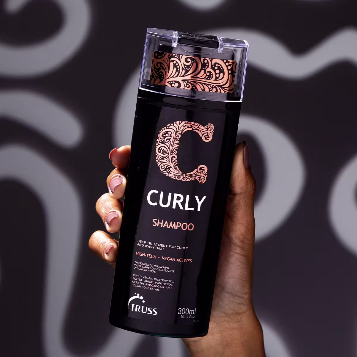 A imagem mostra uma mão feminina segurando a embalagem do Shampoo TRUSS Curly, exibindo o produto. Cores predominantes da embalagem são preto, branco e laranja.