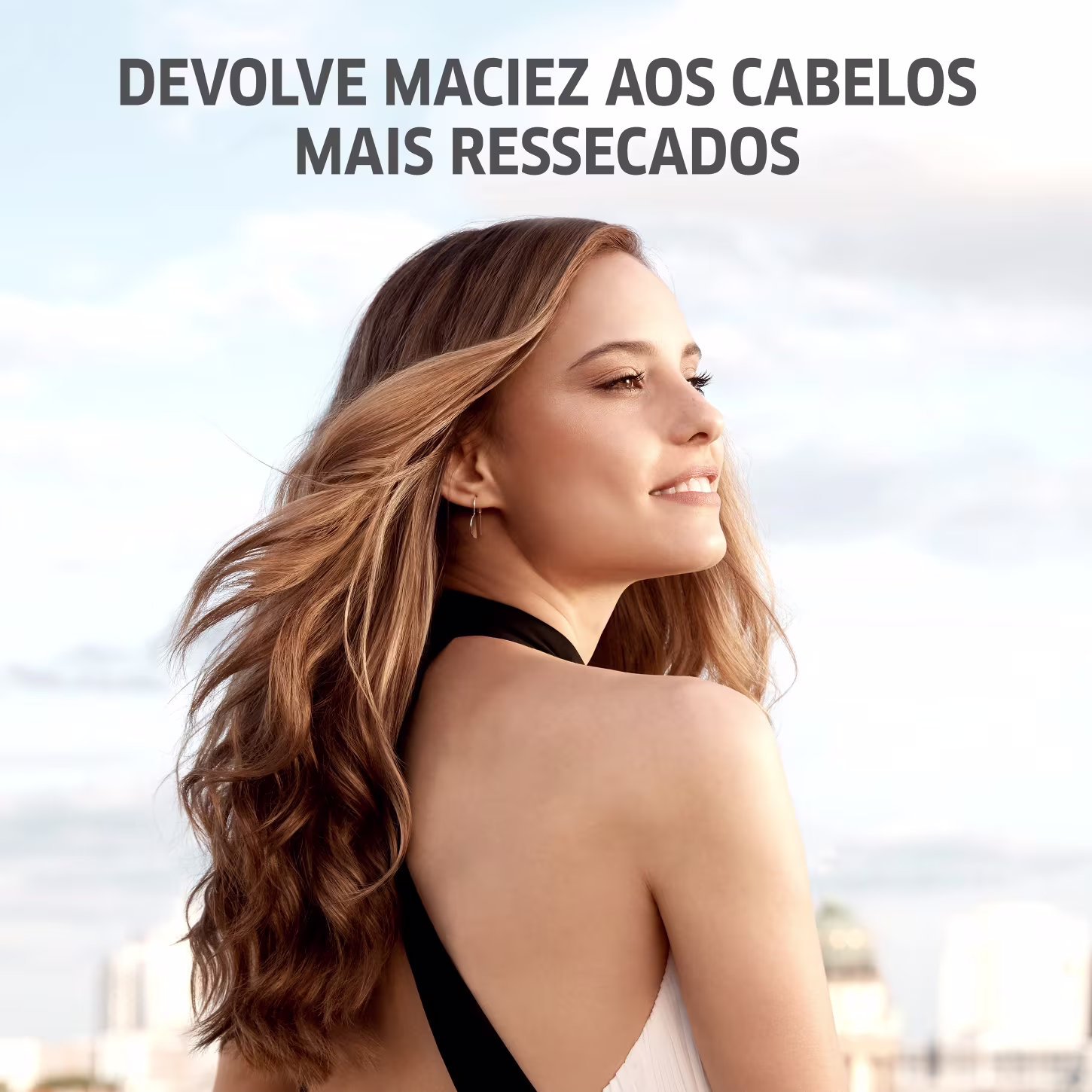 A imagem apresenta uma mulher jovem com longos cabelos ondulados em tons de loiro e castanho. Ela está usando um top preto e tem um sorriso suave no rosto. O texto diz "Devolve maciez aos cabelos mais ressecados".