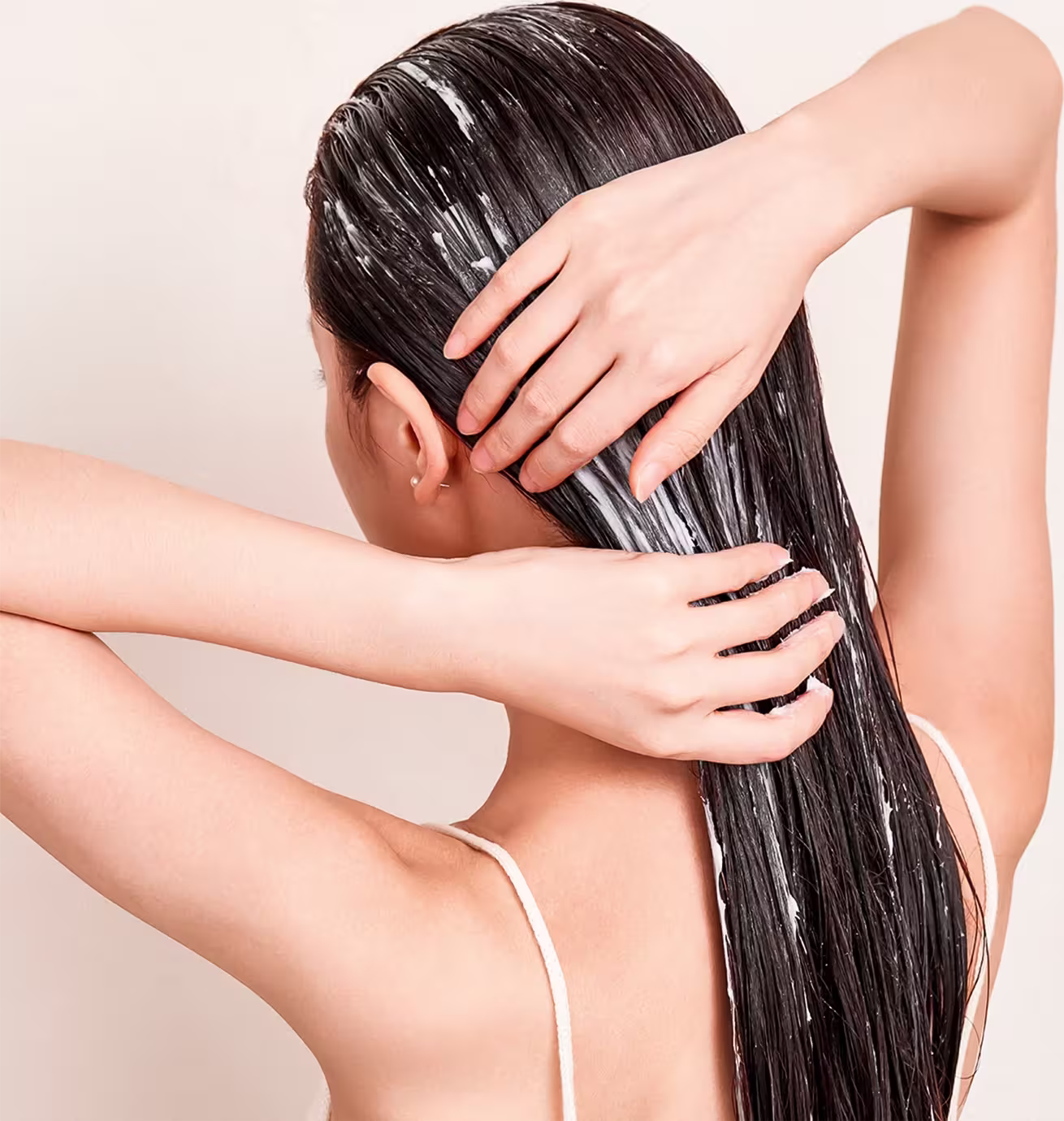 Esta imagem mostra uma mulher com longos cabelos escuros e lisos, aplicando um produto capilar. Suas mãos estão posicionadas em seu couro cabeludo, aparentemente massageando ou aplicando o produto em seus fios.