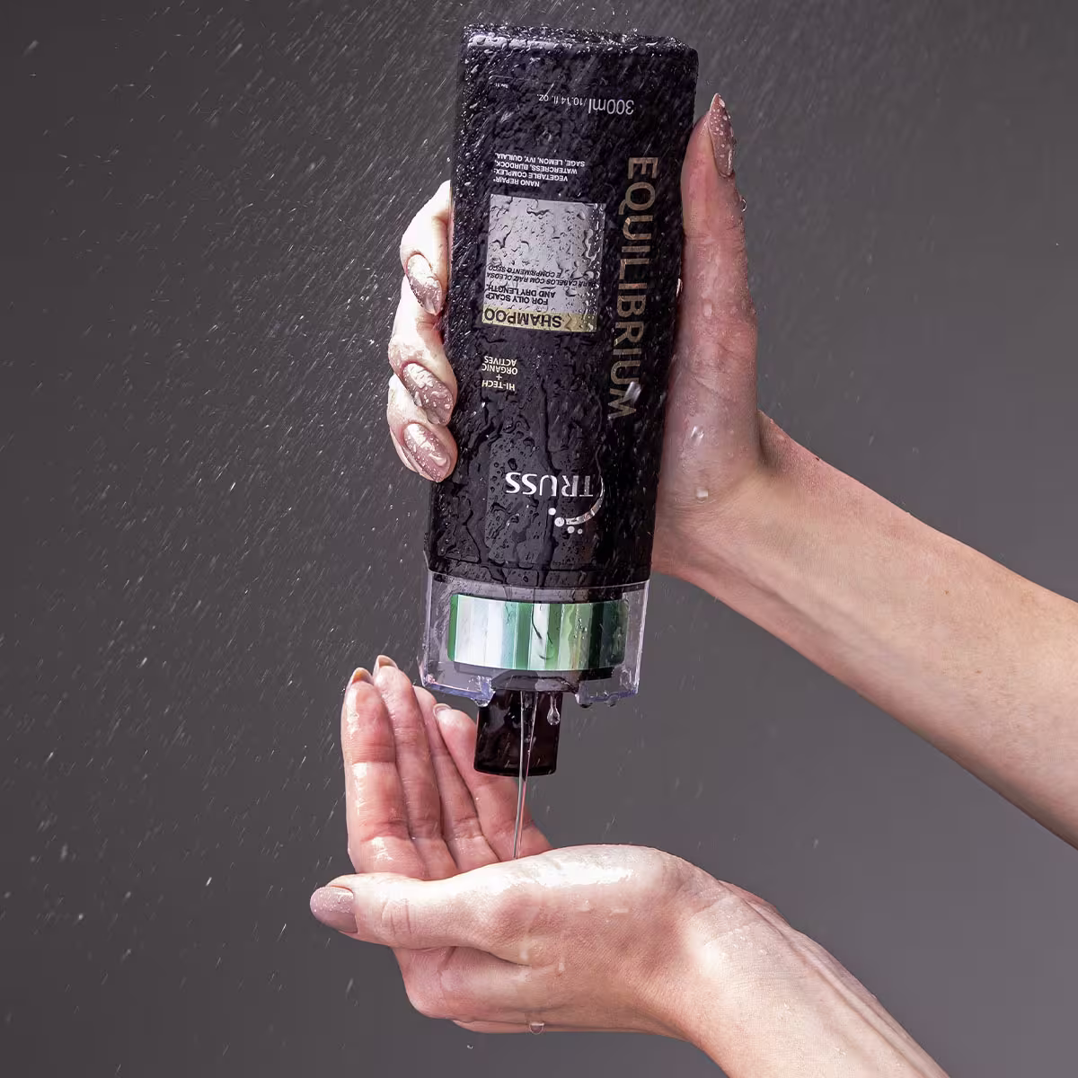 Shampoo "Equilibrium" segurado por uma mão feminina com unhas pintadas, sendo derramado na outra mão. O fundo é cinza, com gotas de água caindo ao redor.