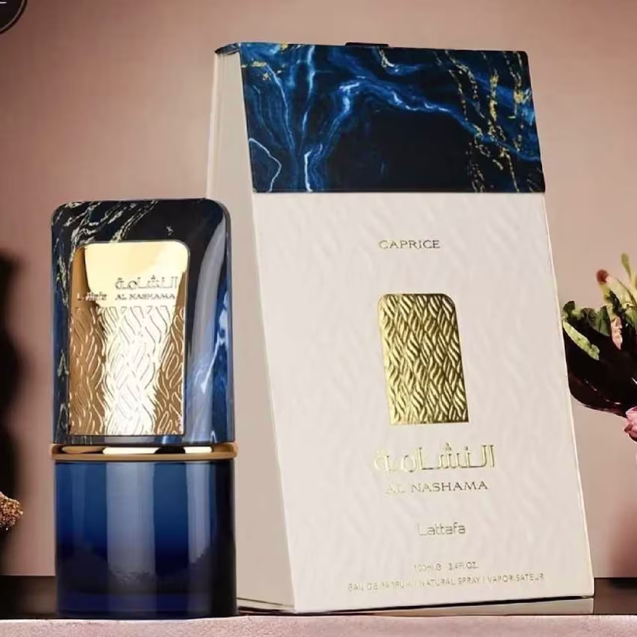 Frasco de perfume azul e branco da marca Lattafa Perfumes, com rótulo dourado e texto em árabe. Caixa de embalagem com padrão azul e branco, exibindo o nome do produto "Al Nashama" em letras douradas.
