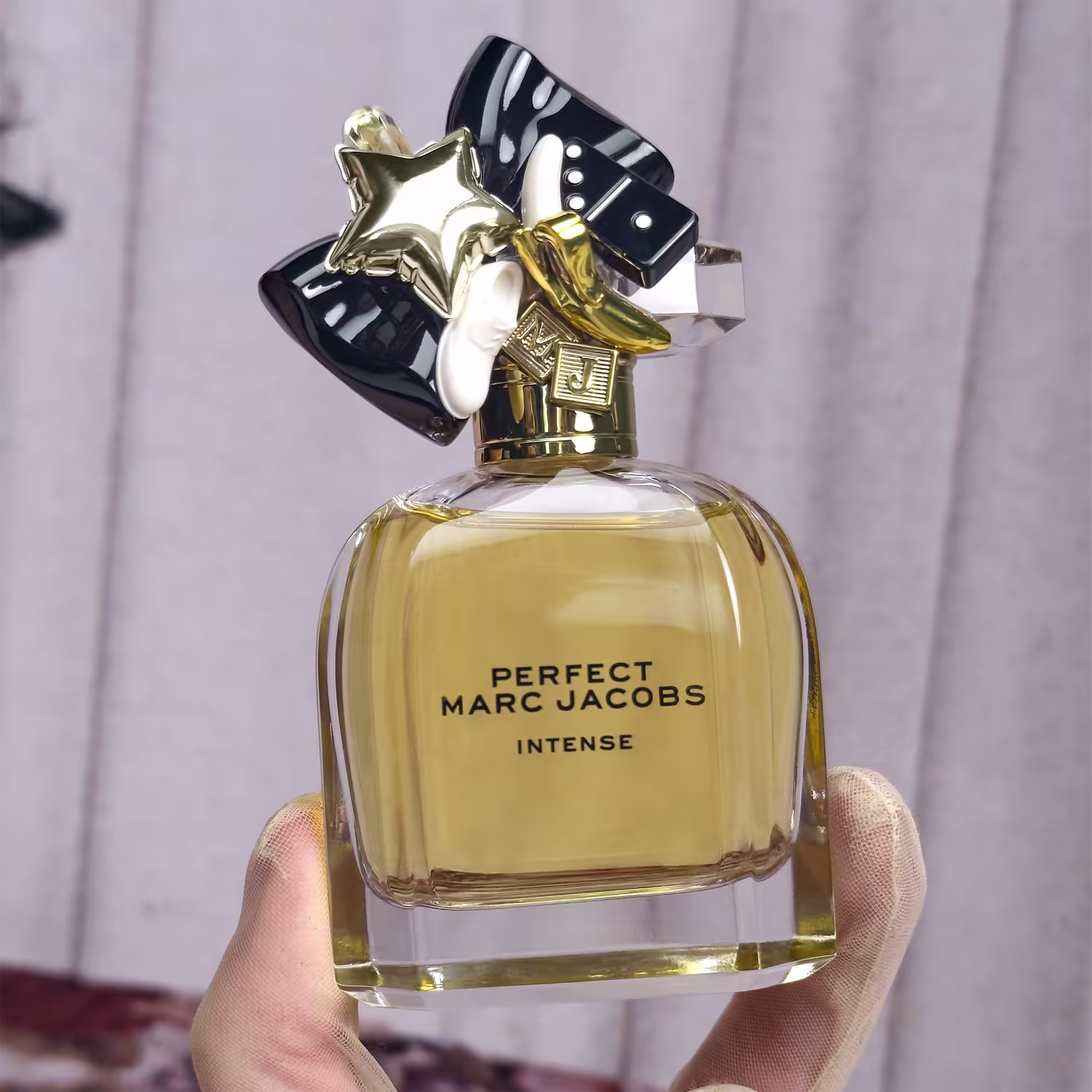 Frasco de perfume dourado e preto da linha "Perfect Marc Jacobs Intense", com tampa decorativa em forma de estrela e laço preto, exibindo o nome da marca e do produto em letras douradas.
