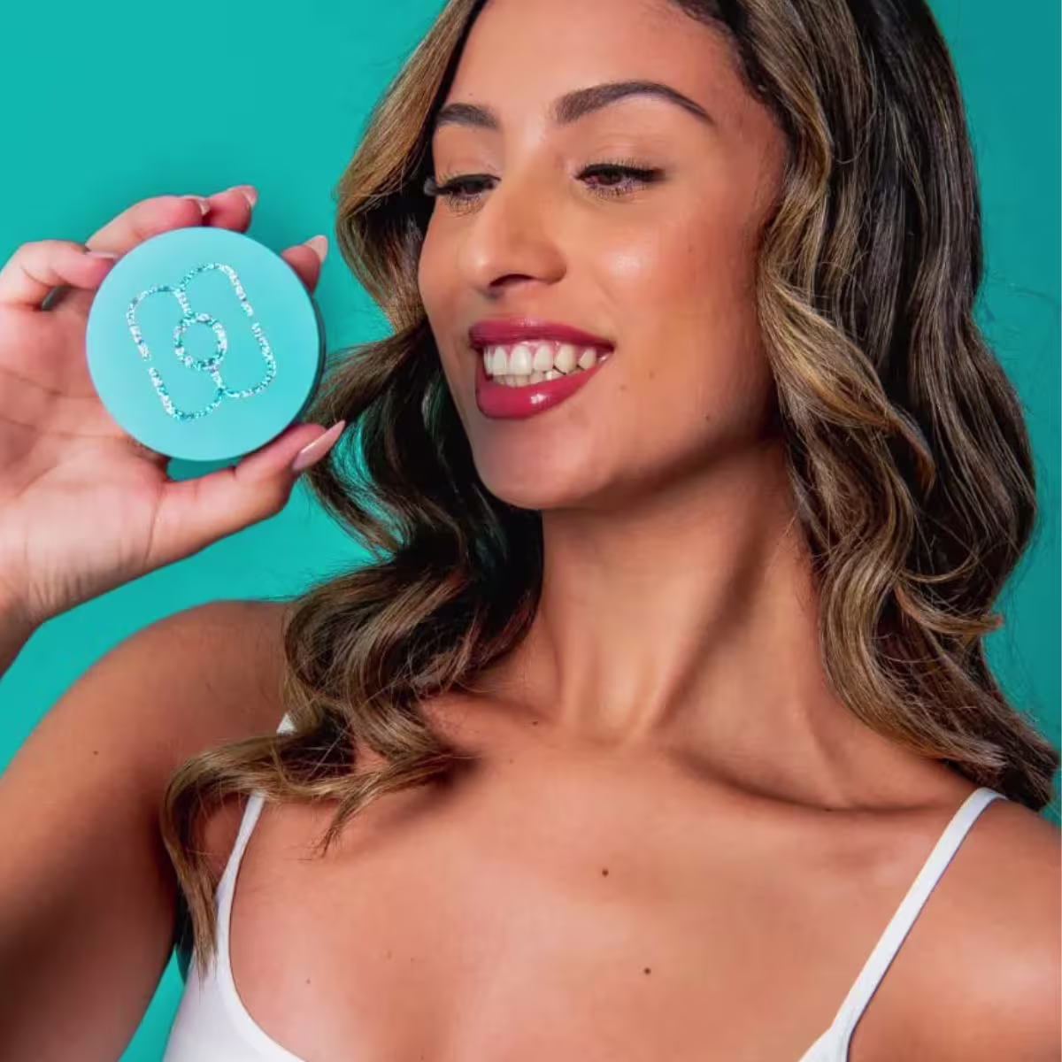 O produto apresentado é um pó compacto de maquiagem facial. A modelo segura o produto próximo ao rosto, exibindo um sorriso com dentes brancos e maquiagem com batom vermelho.