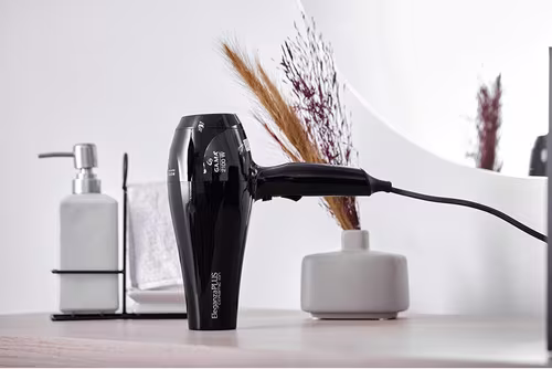 A imagem mostra o secador de cabelo com a ponta do bico apoiada em uma superfície plana. Ao fundo estão hastes de trigo e flores secas decorativas, há também uma embalagem para sabonete líquido ou shampoo e uma toalha branca.