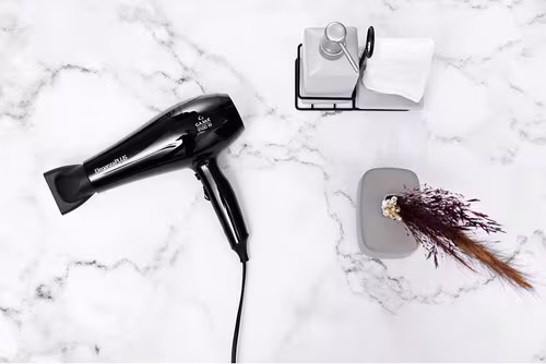 A imagem mostra o secador de cabelo sobre uma superfície que parece mármore. Ao lado dele há uma embalagem para colocar shampoo, sabonete líquido ou outro item de higiene. Há também um frasco cinza com algumas folhas em tom terroso.