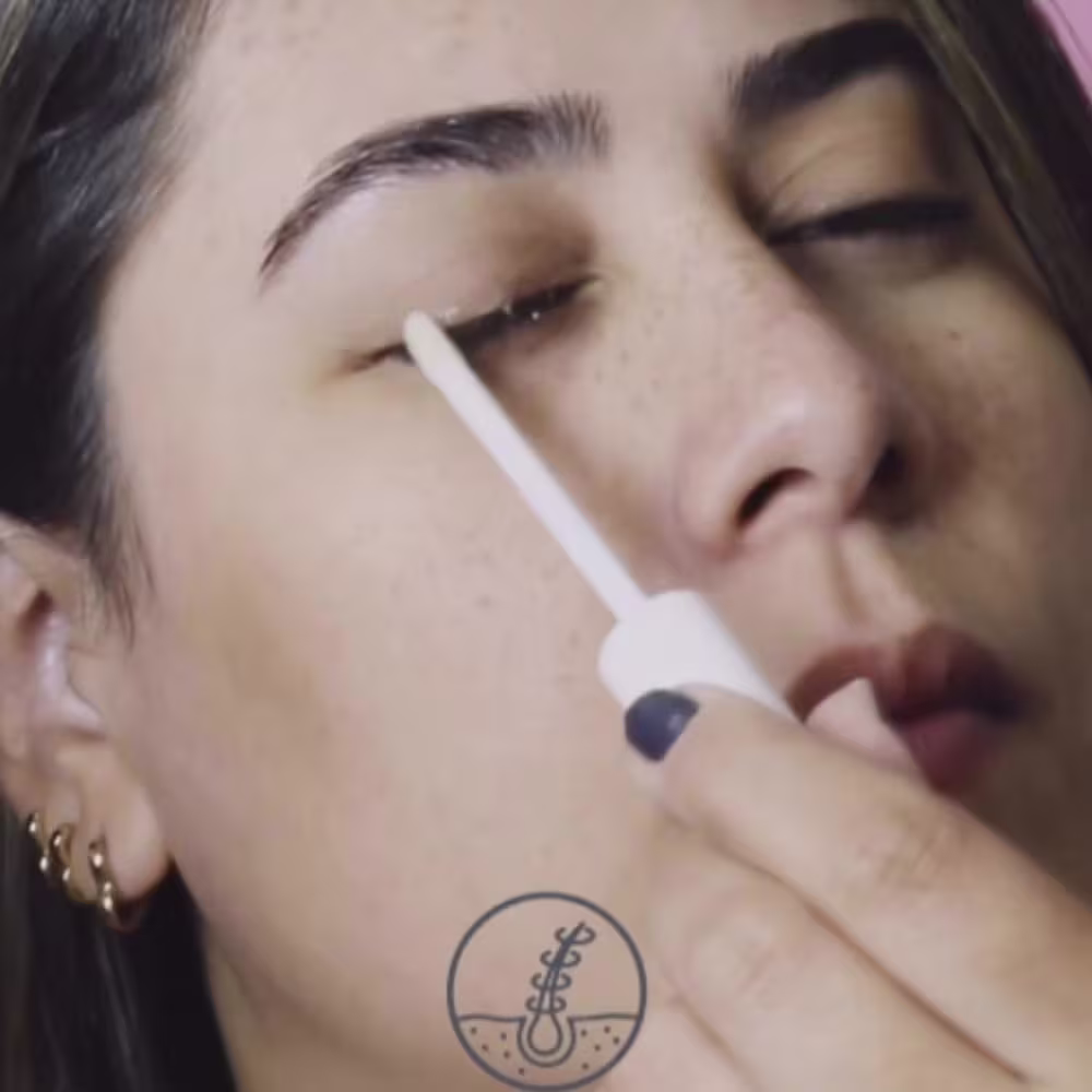 A pessoa está aplicando o  produto nos cílios. O ambiente sugere um contexto de cuidados pessoais e beleza.