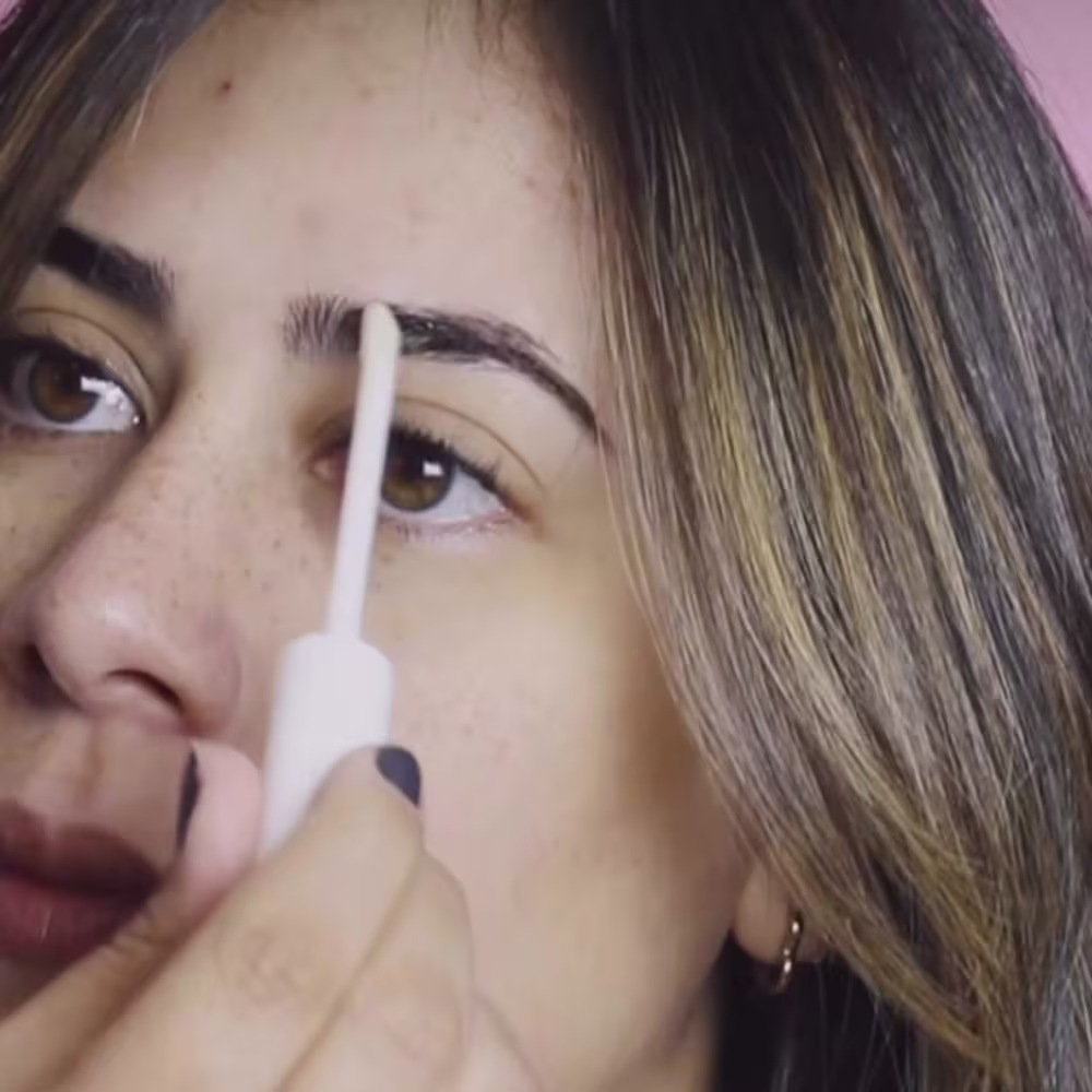 A imagem mostra uma mulher aplicando o produto em suas sobrancelhas, em um ambiente iluminado e com tons neutros predominantes.