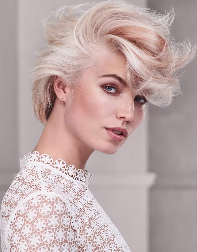 Esta imagem apresenta uma modelo feminina com cabelos platinados e estilo despojado. Ela está usando uma blusa rendada branca com detalhes em formato de flores vazadas.