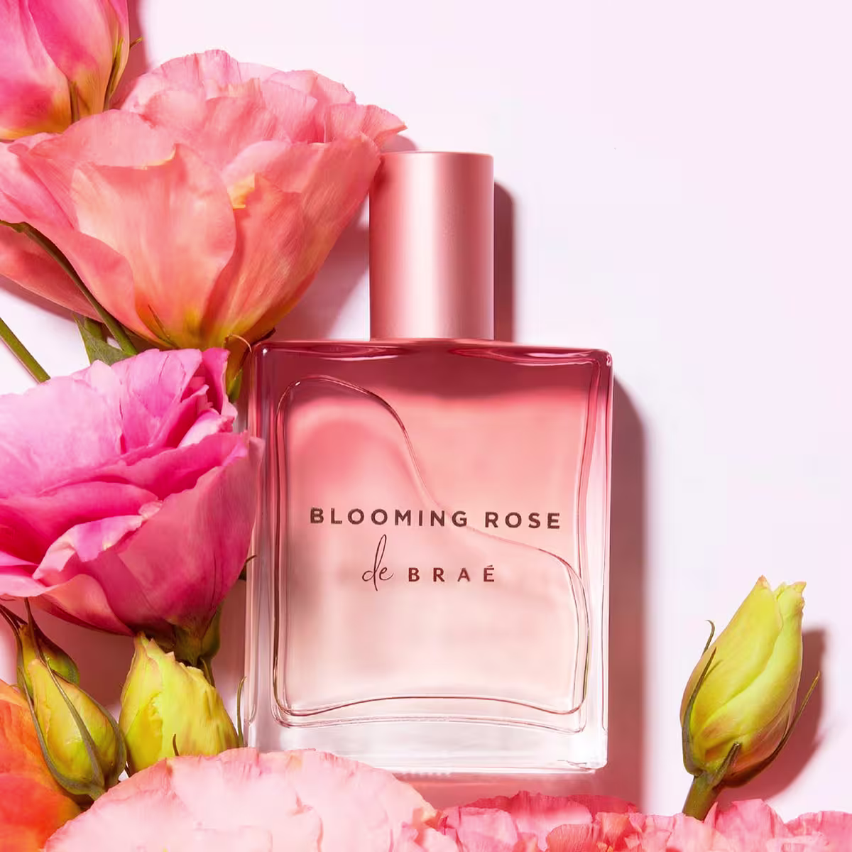 Perfume de cor rosa metálico em frasco retangular de vidro transparente. A embalagem tem um rótulo com o texto "BLOOMING ROSE de BRAÉ" em letras pretas. O frasco está cercado por flores rosas, amarelas e laranjas, aparentando serem rosas e cravos.