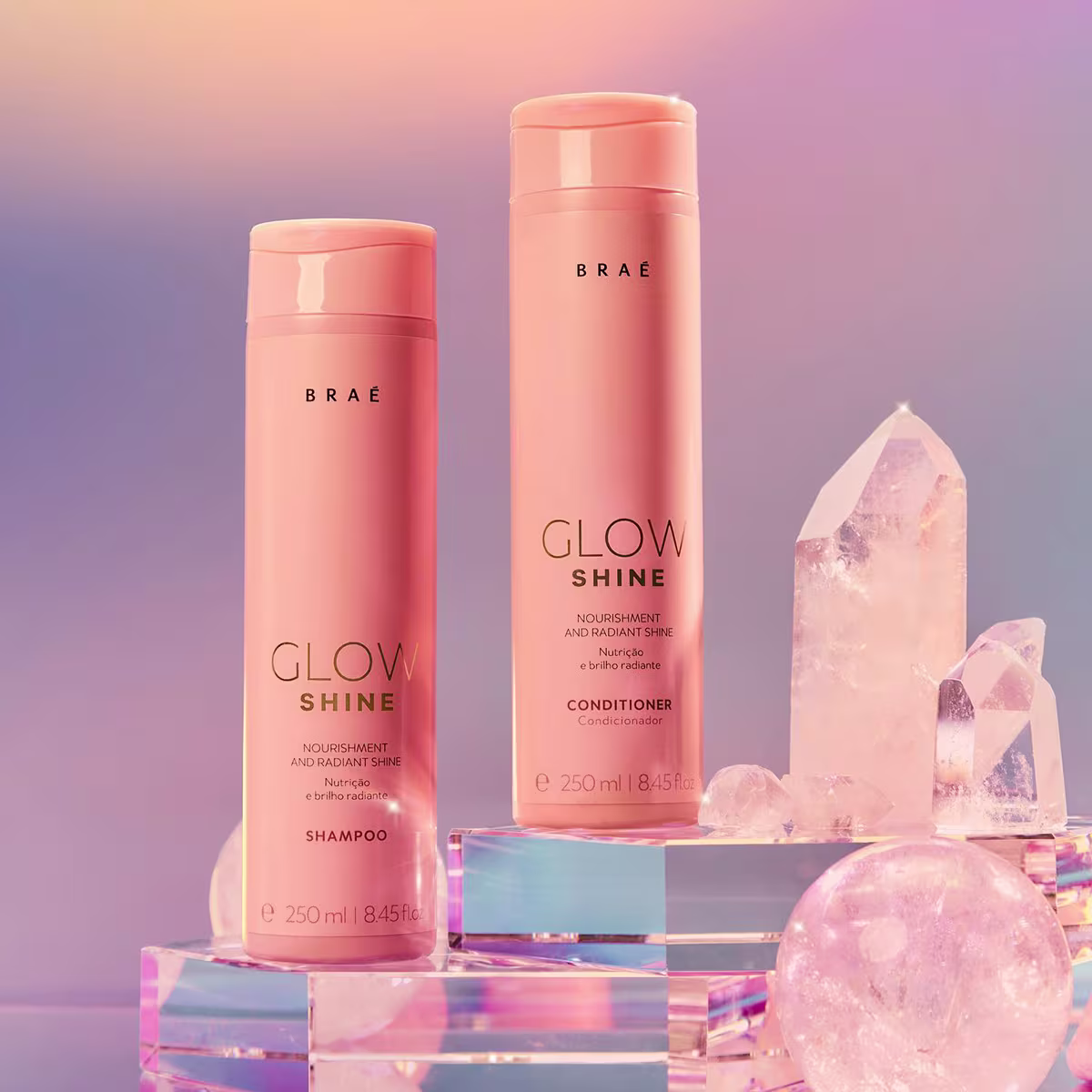 Dois frascos de shampoo e condicionador da linha Glow Shine da marca Brae. Os frascos cilíndricos são de plástico na cor pêssego. Ao lado, há cristais de quartzo rosa em diferentes formatos e tamanhos, refletindo a luz.