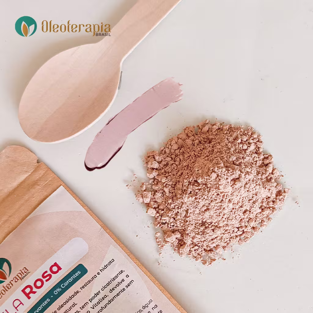 A imagem mostra uma superfície branca e sobre ela está: a embalagem da argila rosa, uma colher de madeira pequena, uma amostra da argila úmida pronta e uma porção pequena da argila em pó.