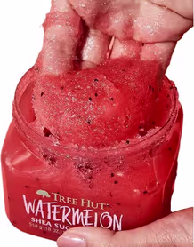 Produto de esfoliação corporal Tree Hut Watermelon Shea Sugar Scrub. Embalagem transparente com tampa laranja brilhante. O frasco contém um esfoliante de açúcar de cor rosa vibrante com pequenas sementes escuras visíveis.