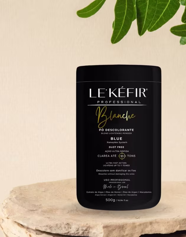 A imagem mostra a embalagem preta do pó descolorante posicionada sobre uma pedra clara, em um fundo bege, com folhas verdes decorando a cena, transmitindo uma atmosfera natural para esse cosmético capilar.