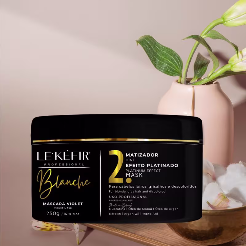 A imagem mostra a embalagem da máscara capilar posicionada ao lado de uma vasilha dourada com plantas e flores brancas, criando um cenário natural e elegante para apresentar o produto. As flores parecem ser orquídeas, com suas folhas verdes e hastes.