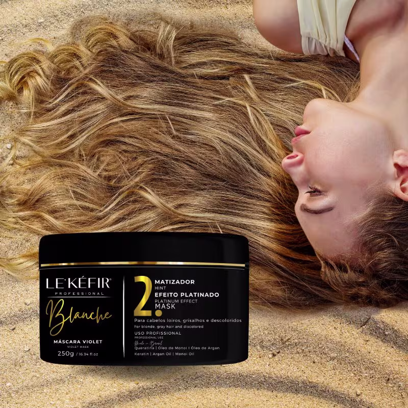 A imagem mostra o pote da máscara capilar posicionada junto a longos cabelos loiros espalhados, pertencentes a uma pessoa que não é totalmente visível na foto.