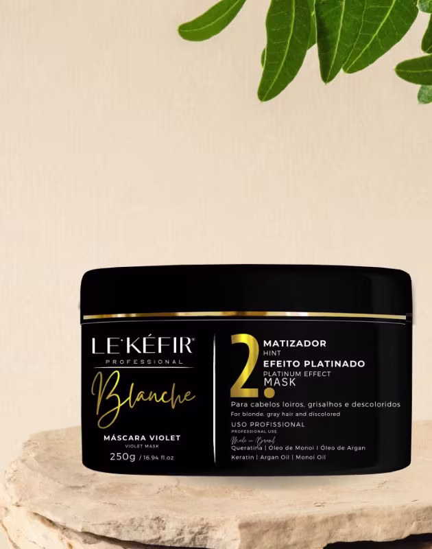 A imagem mostra a embalagem preta da máscara capilar posicionada sobre uma pedra clara, em um fundo bege, com folhas verdes decorando a cena, transmitindo uma atmosfera natural para esse cosmético capilar.