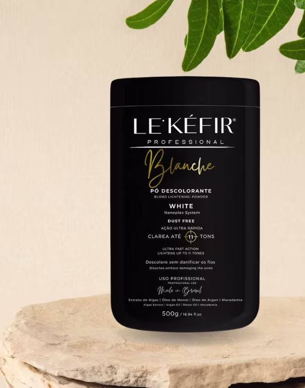 A imagem mostra a embalagem preta do pó descolorante posicionada sobre uma pedra clara, em um fundo bege, com folhas verdes decorando a cena, transmitindo uma atmosfera natural para esse cosmético capilar.