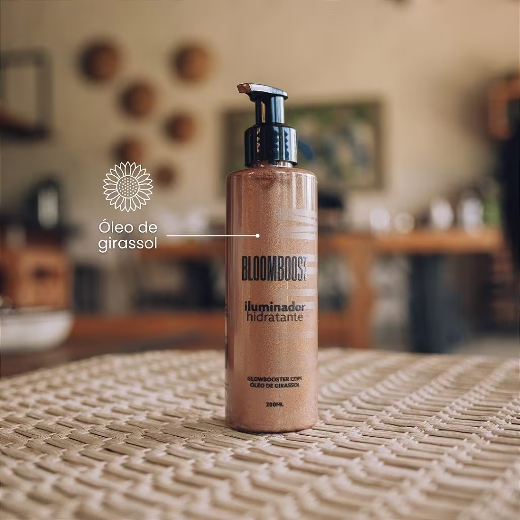 Frasco do iluminador corporal, com uma cor marrom clara, posicionado sobre um tapete trançado em um ambiente decorado com peças de barro ou cerâmica penduradas ao fundo. A foto menciona que contém óleo de girassol como um dos ingredientes principais.