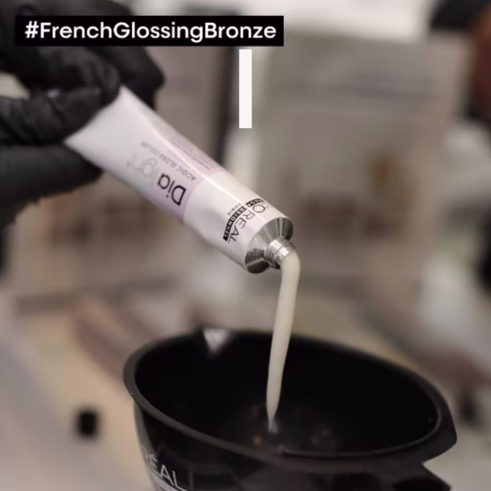 O produto mostrado é um tubo de tonalizante da marca L'Oreal Professionnel. O tubo é branco com o nome do produto e da linha impressos em preto. O creme está sendo dispensado do tubo em um recipiente preto, formando um fluxo cremoso.