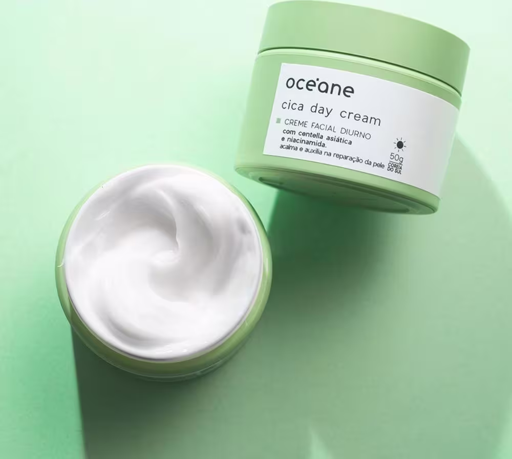 A imagem mostra 2 potes do Océane Cica Day Cream vistos de cima sobre um fundo verde. Um deles está aberto e é possível ver o conteúdo branco e cremoso dentro, O outro está deitado de maneira a focar no rótulo.