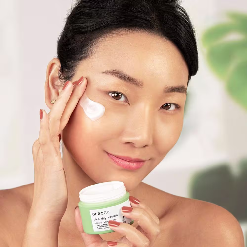 A imagem mostra uma mulher aparentemente asiática sorrindo enquanto segura o Cica Day Cream em uma mão e aplica o creme no rosto com a outra.