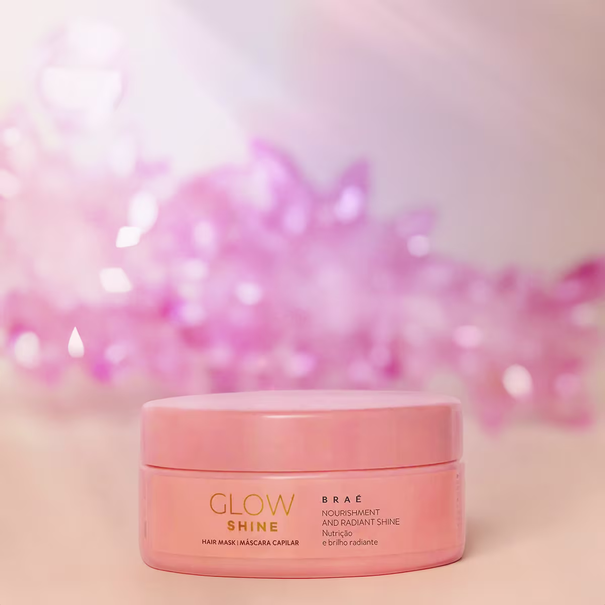 Máscara capilar Glow Shine da linha Braé. Embalagem redonda de plástico na cor rosa claro. A embalagem apresenta tampa removível na mesma tonalidade. Ao fundo aparecem cristais rosados desfocados.
