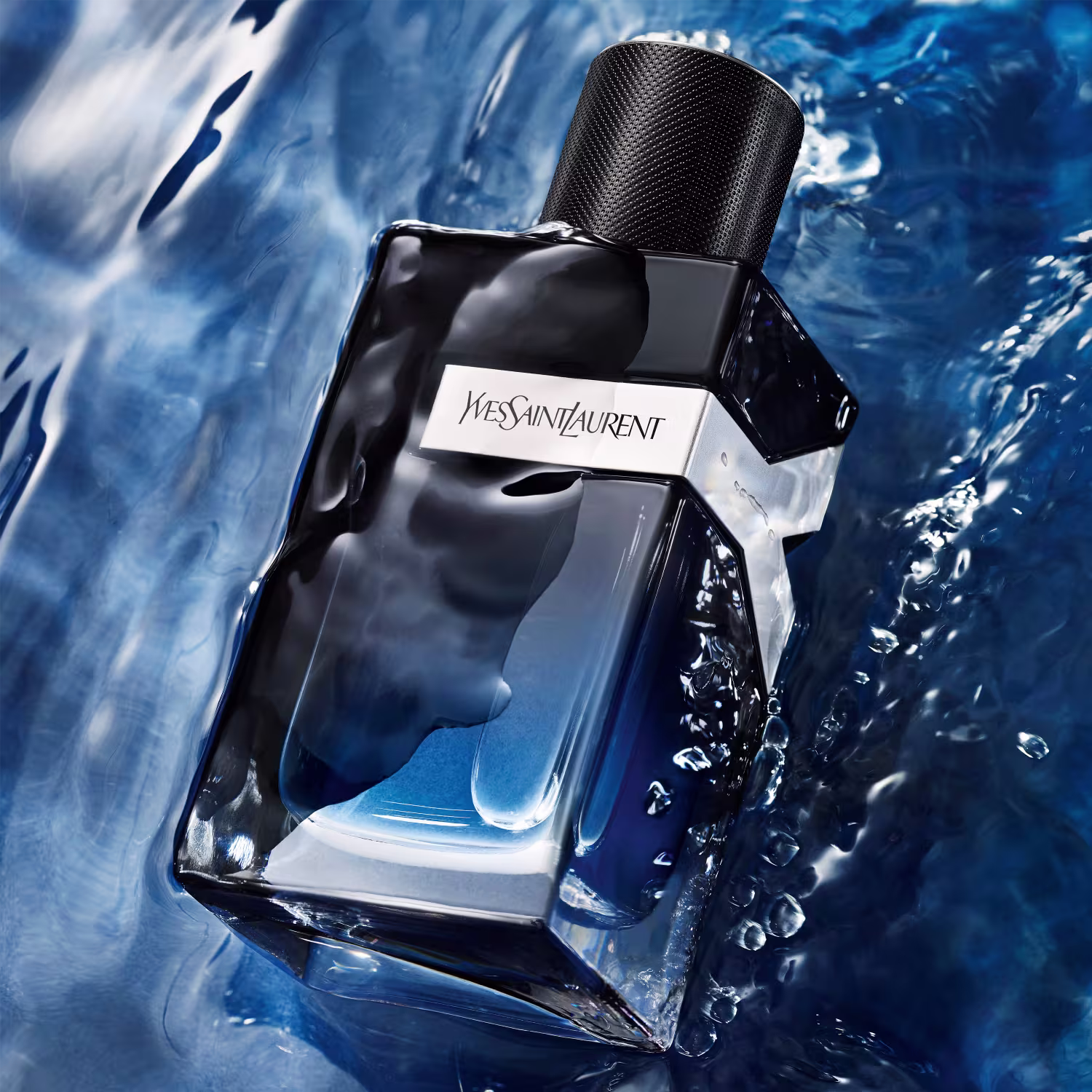 Perfume em frasco retangular de vidro preto com detalhes em azul claro. Rótulo branco com o nome "Yves Saint Laurent" impresso. O frasco está parcialmente submerso em água azul com respingos e bolhas ao redor, criando um efeito refrescante.