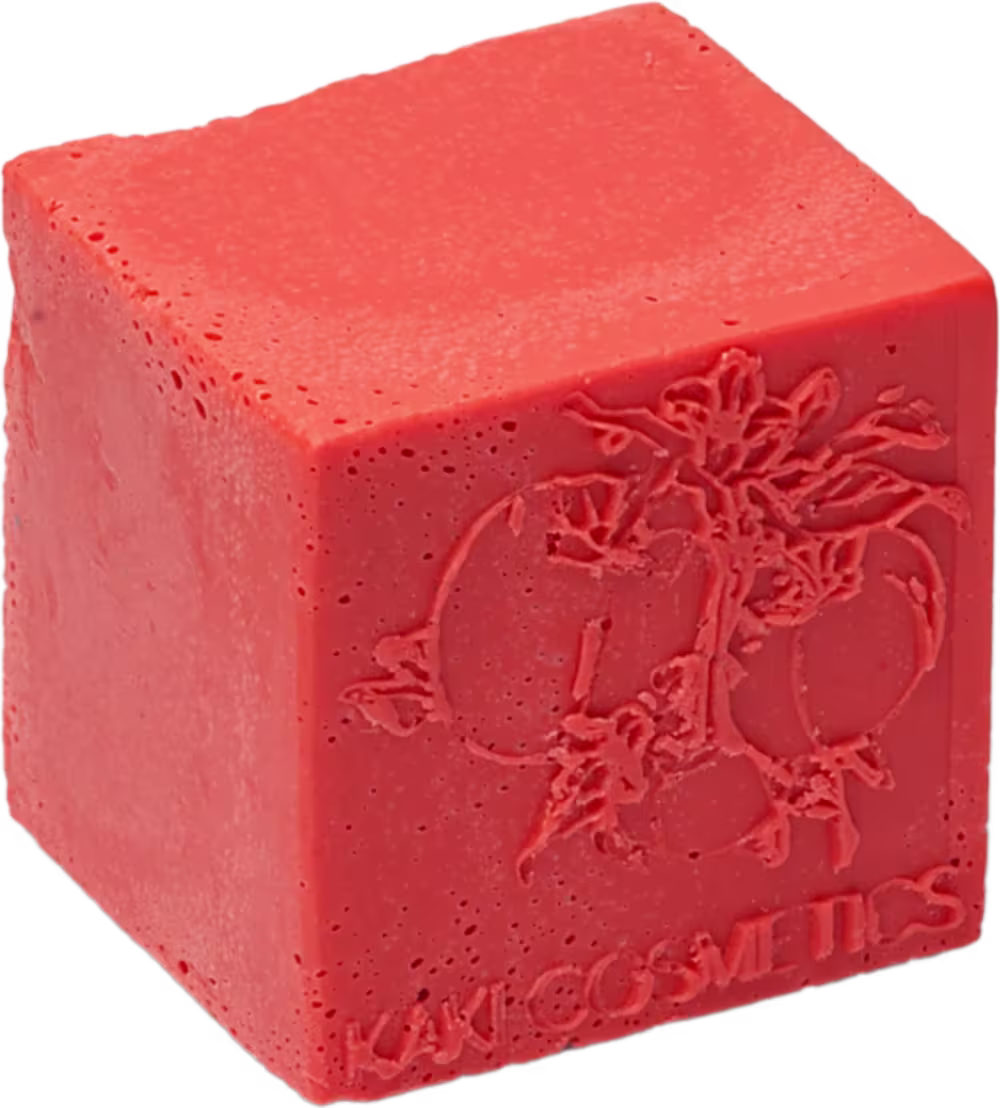 Barra de condicionador em formato cúbico, de cor vermelho-alaranjado. A superfície apresenta uma textura levemente porosa. No centro, há um emblema em relevo representando uma flor estilizada.