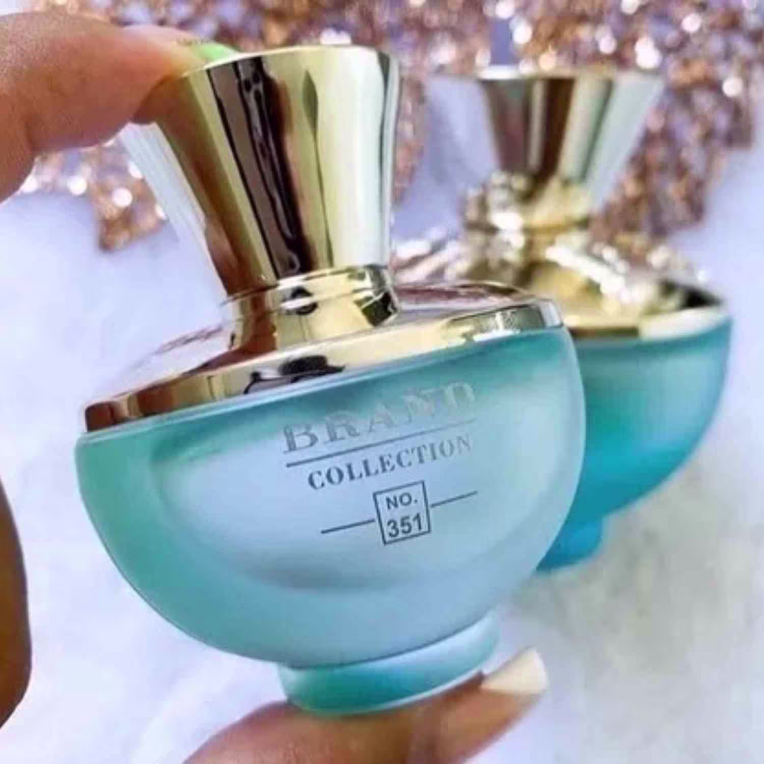 A imagem mostra uma mão feminina segurando o frasco do perfume. Ao fundo há um fundo branco com detalhes dourados e outro frasco de perfume.