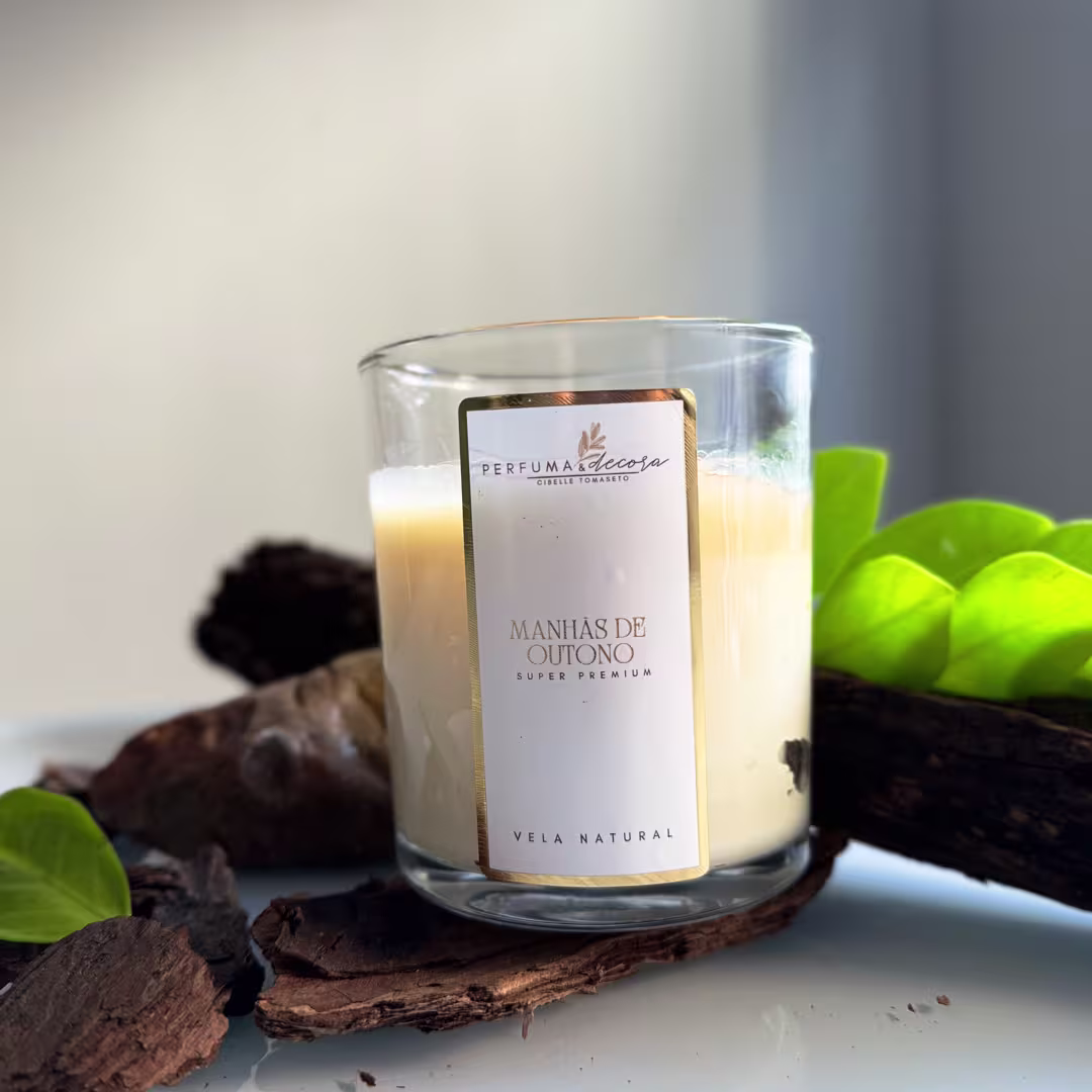 Vela natural em frasco de vidro transparente. O frasco tem uma etiqueta branca com detalhes em dourado que diz "Perfuma & Decora Manhãs de Outono". Ao redor do frasco, há folhas verdes e pedaços de casca de árvore, criando um ambiente outonal.