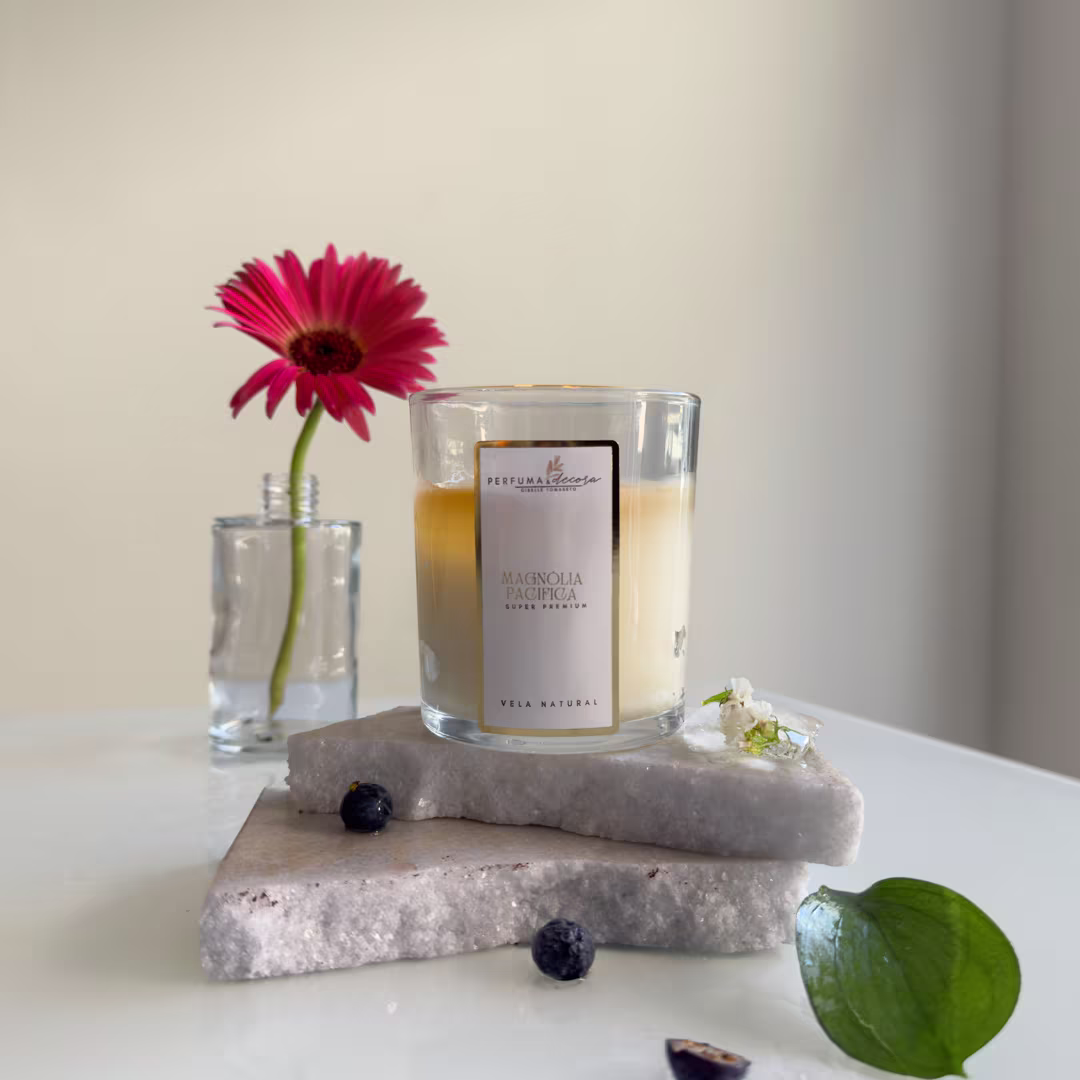 Vela natural em vidro transparente. Rótulo "Perfuma & Decora" e texto "Magnolia Pacifica" na embalagem. Uma flor gerbera vermelha em um pequeno vaso de vidro ao lado. Pedras cinza, folha verde e flores brancas pequenas complementam a composição.